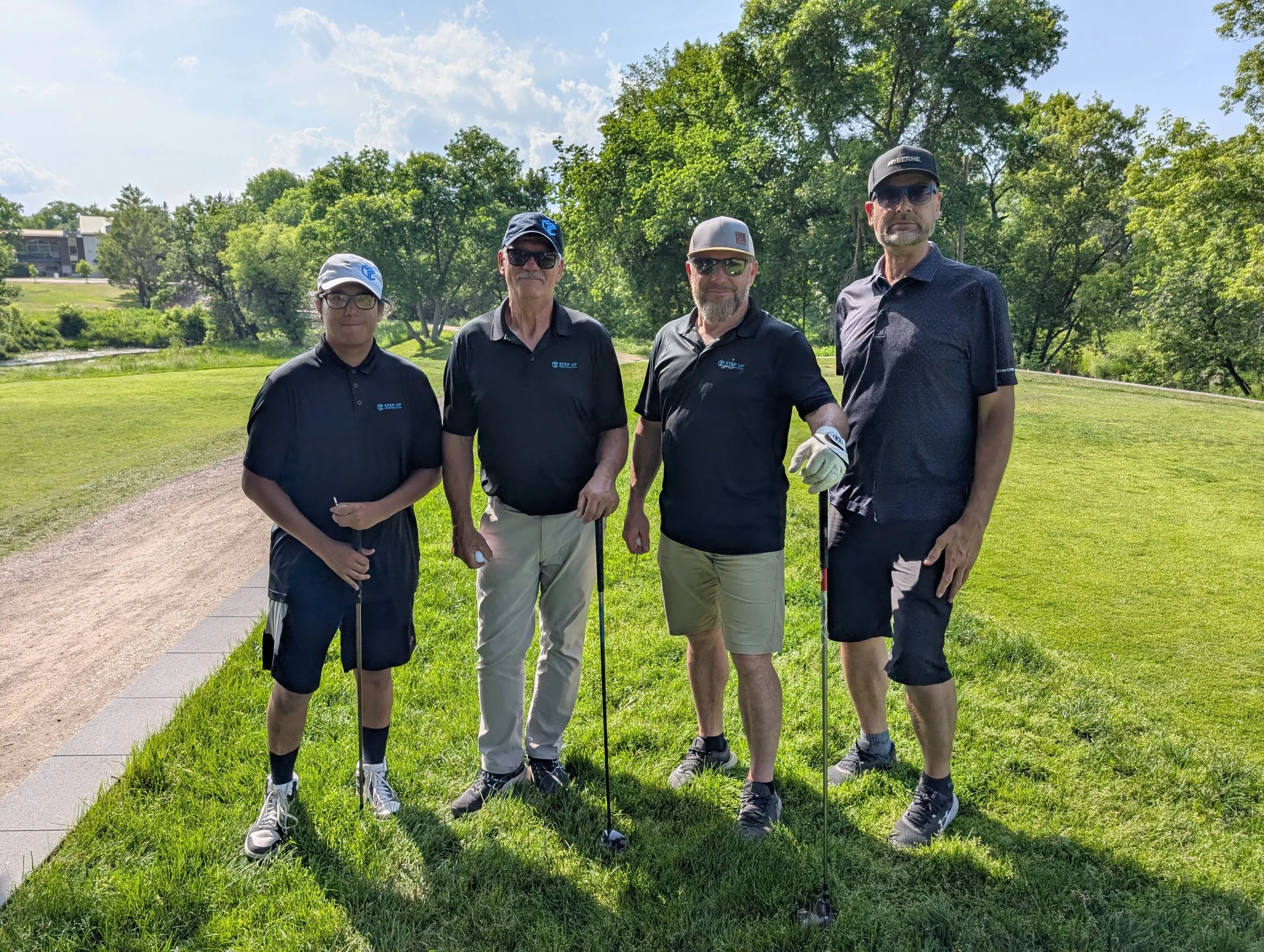 Four men standing on a golf course, holding golf clubs, wearing black shirts, hats, and sunglasses on a sunny day with green trees in the background.
