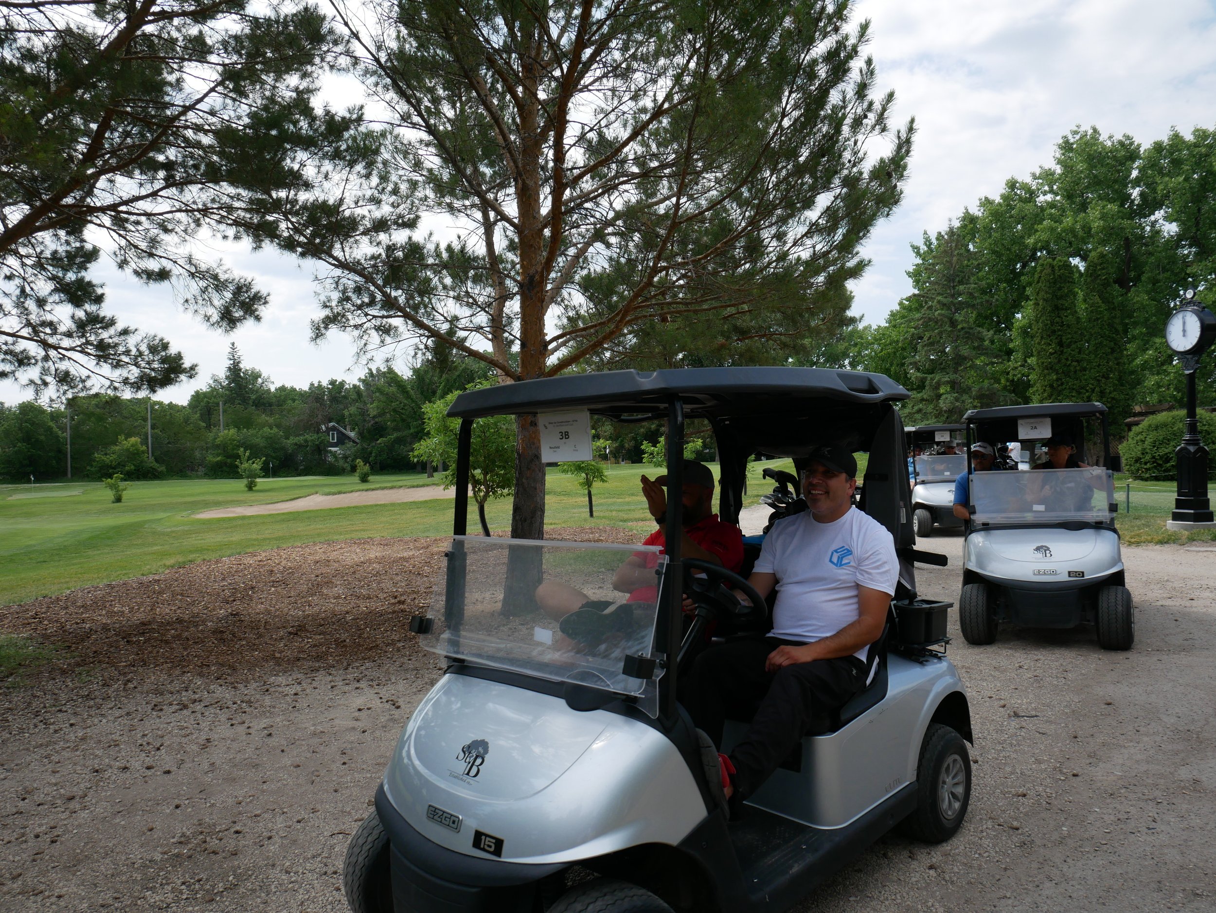 Two men riding in a golf cart on a golf course with trees and a clock in the background.