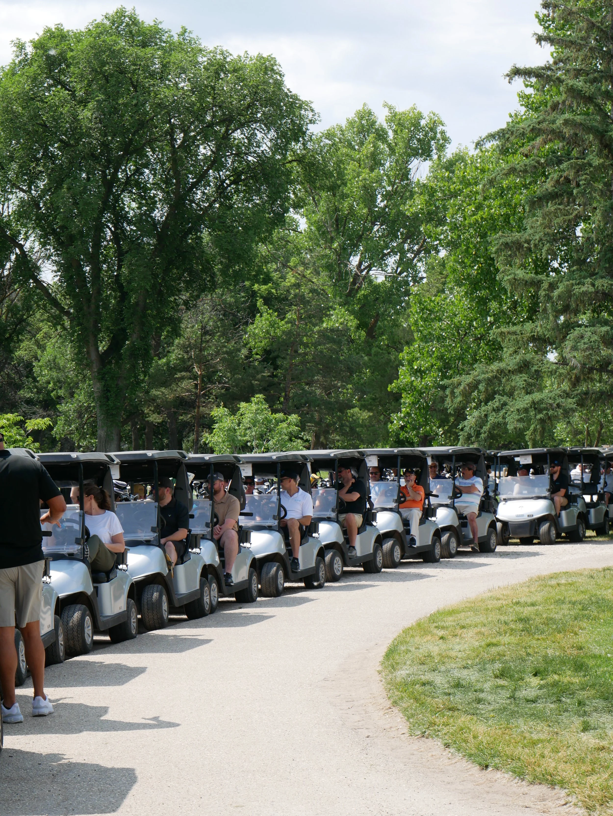 A line of people seated in golf carts on a gravel path in a park or golf course, with tall green trees and a partly cloudy sky in the background.
