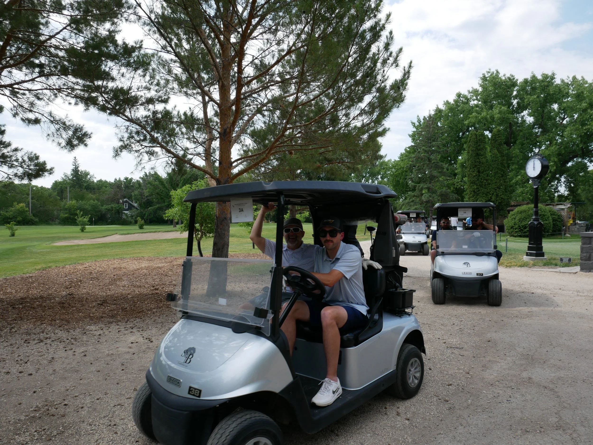 A group of people riding in multiple golf carts on a golf course with trees and a clock in the background.