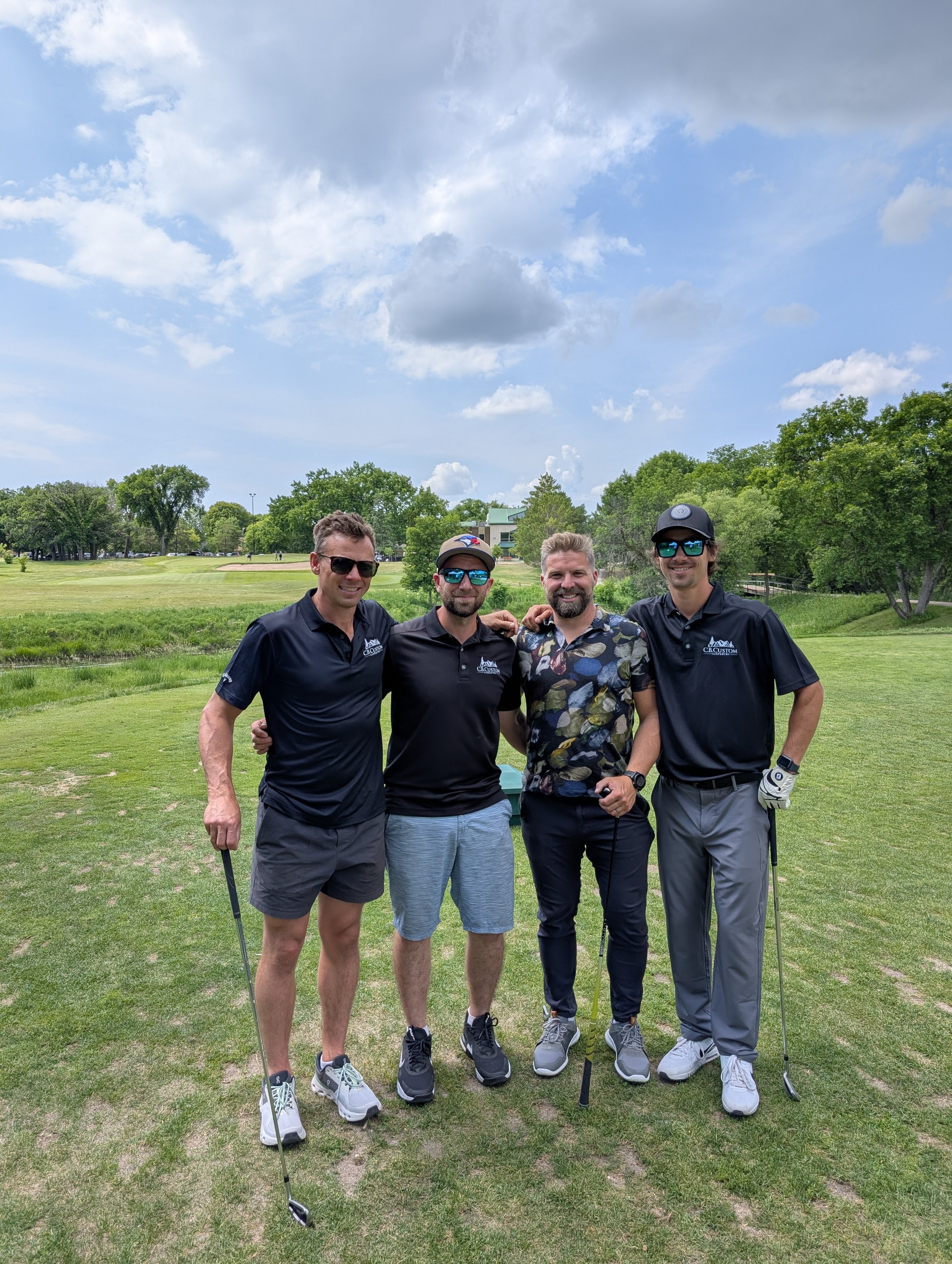 Four men standing on a golf course, smiling and posing with golf clubs, under a partly cloudy sky.