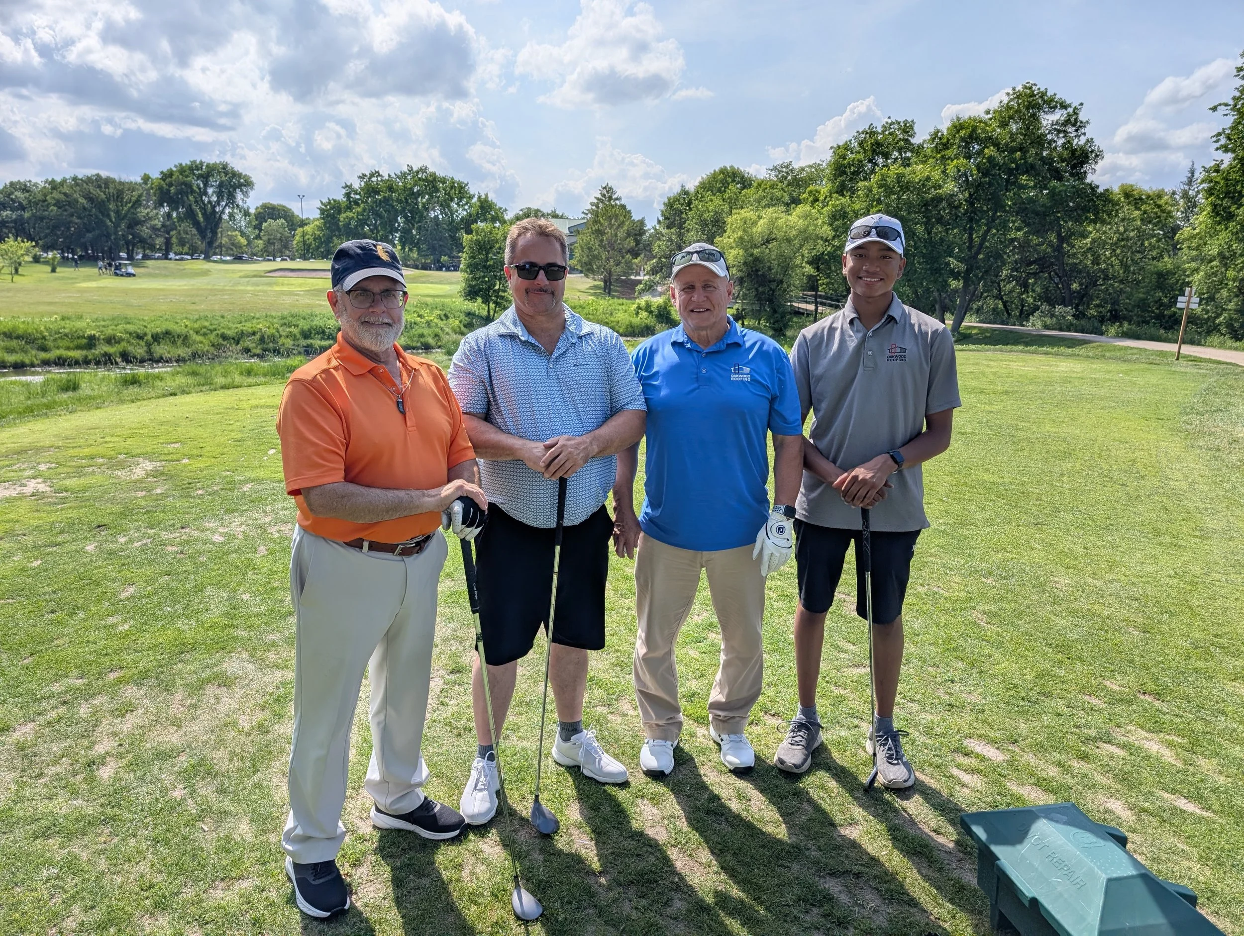 Four men standing on a golf course holding golf clubs, with green grass, trees, and a partly cloudy sky in the background.