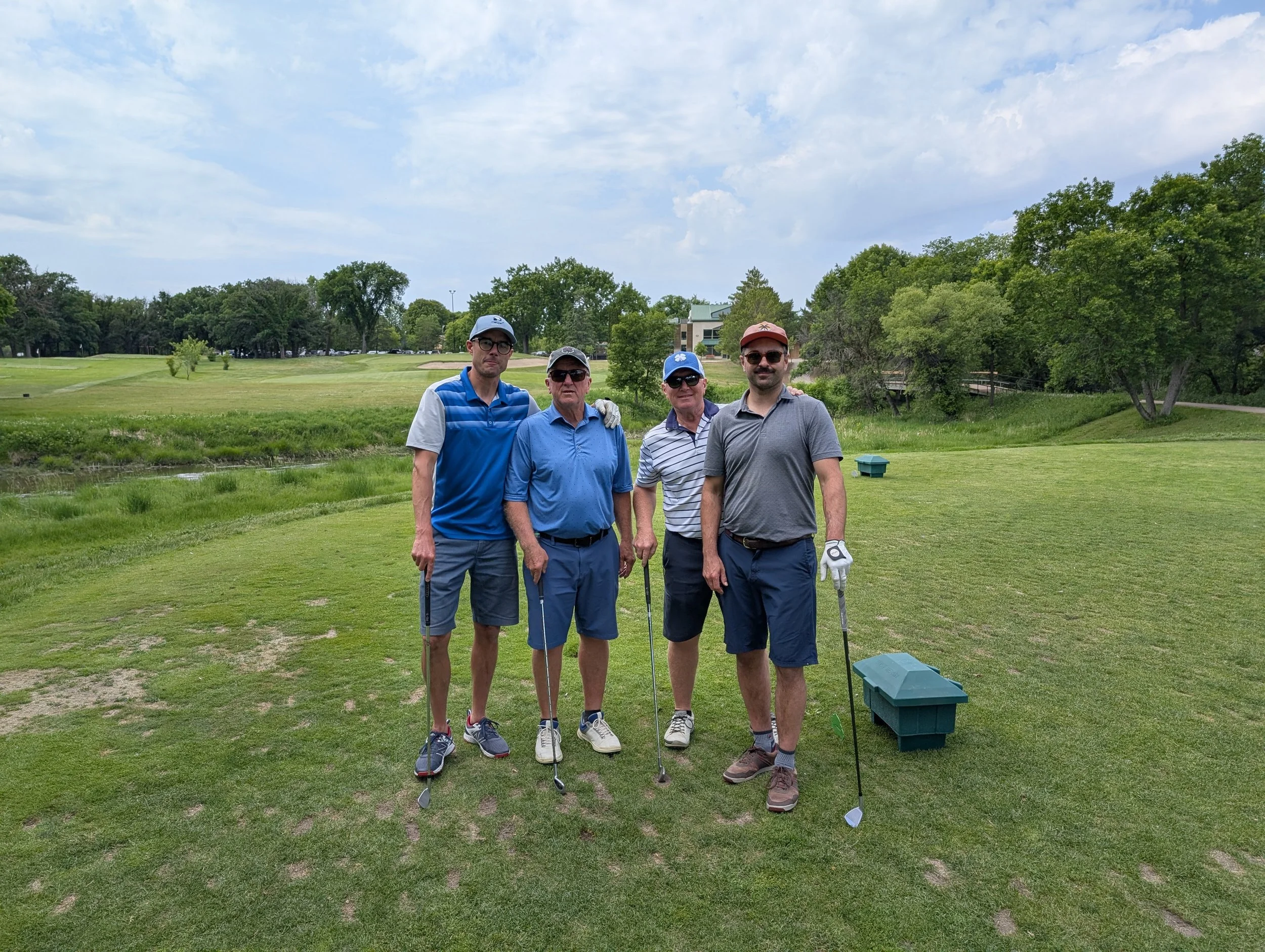 Four men standing on a golf course holding golf clubs, with green grass, trees, a small pond, and a clubhouse in the background.