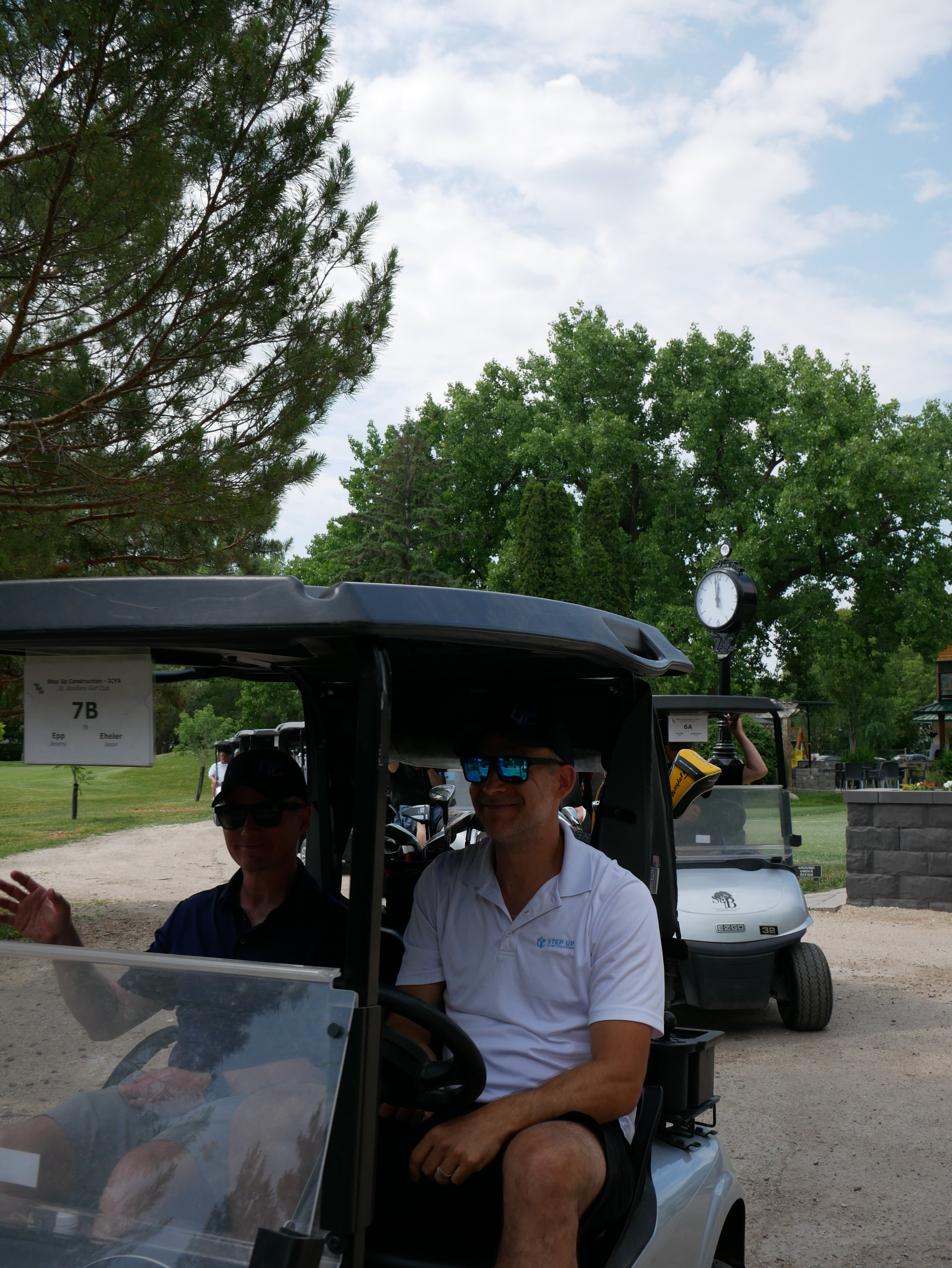Two men sitting in a golf cart on a golf course with green trees and a clock tower in the background.
