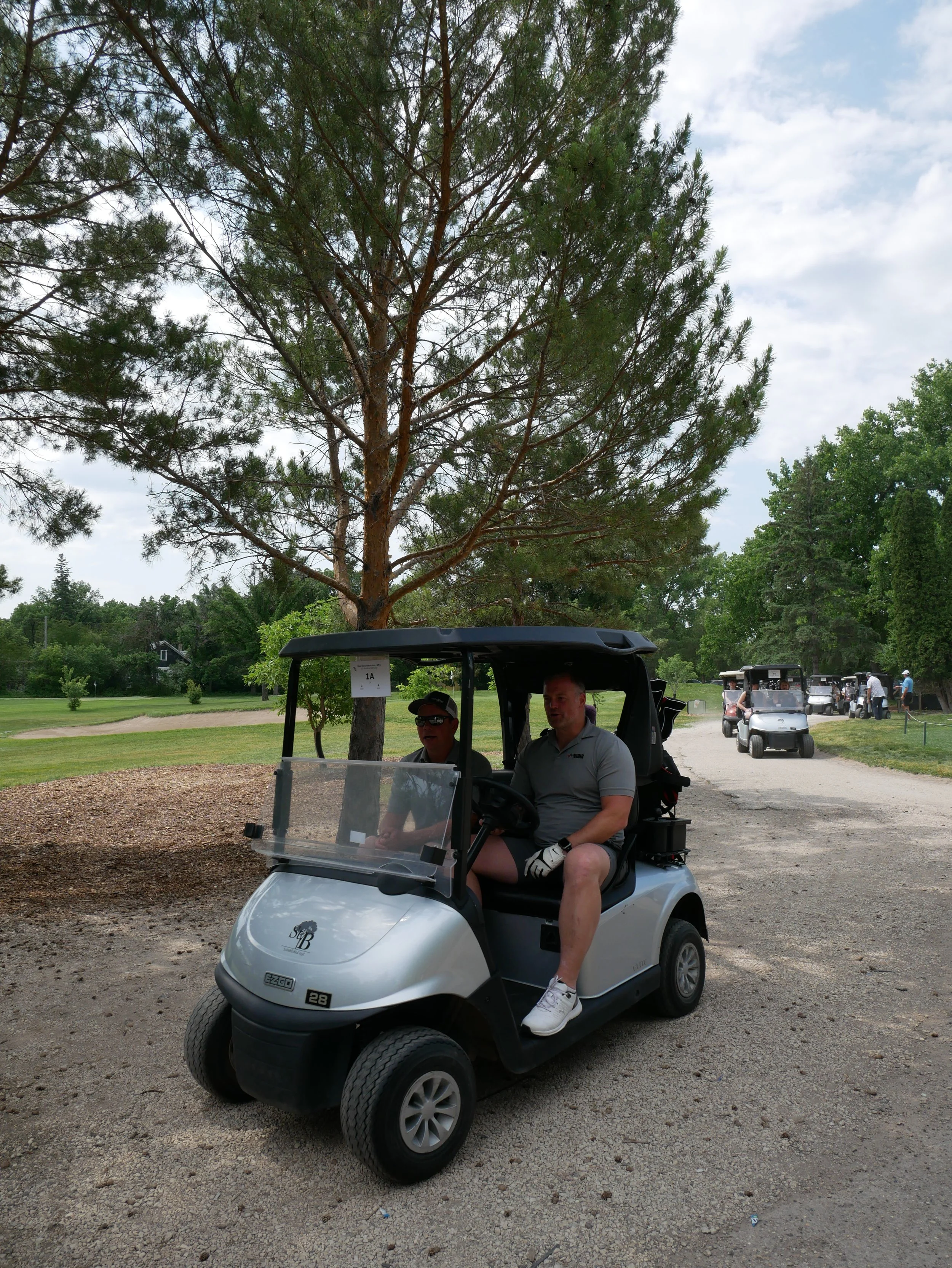 Two men sitting in a golf cart on a golf course, with trees and other golf carts in the background.