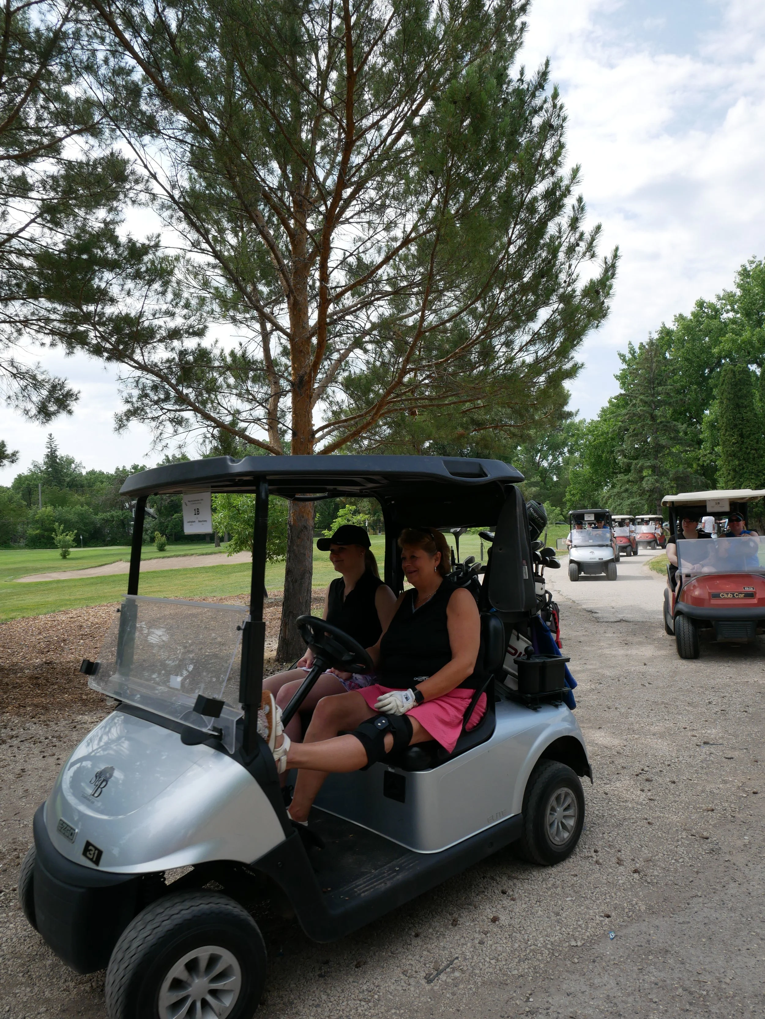 Two women sitting in a golf cart at a golf course, with several other golf carts in the background, trees, and partly cloudy sky.