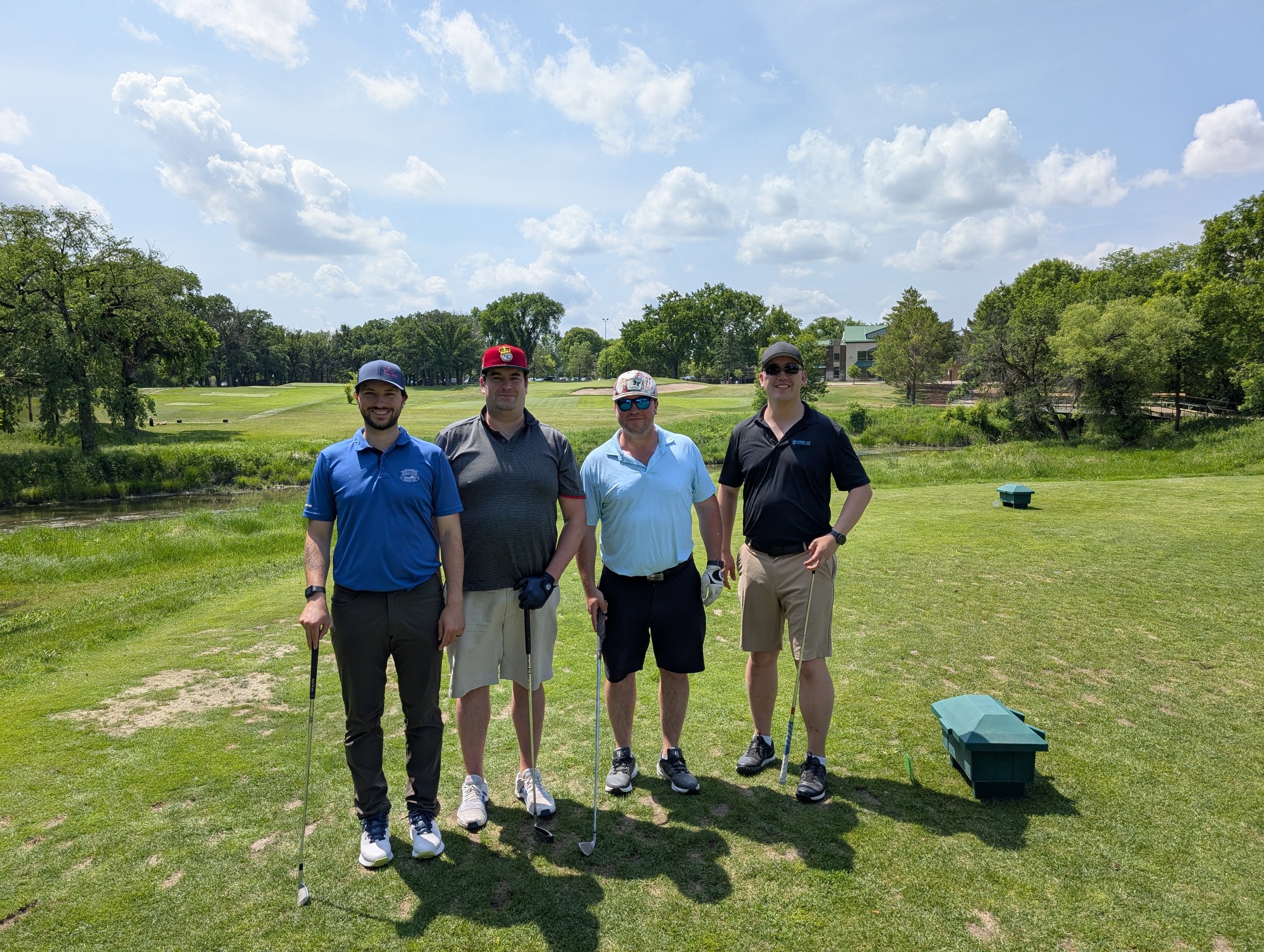 Four men standing on a golf course holding golf clubs, wearing casual golf attire and sunglasses on a sunny day with green trees and a building in the background.