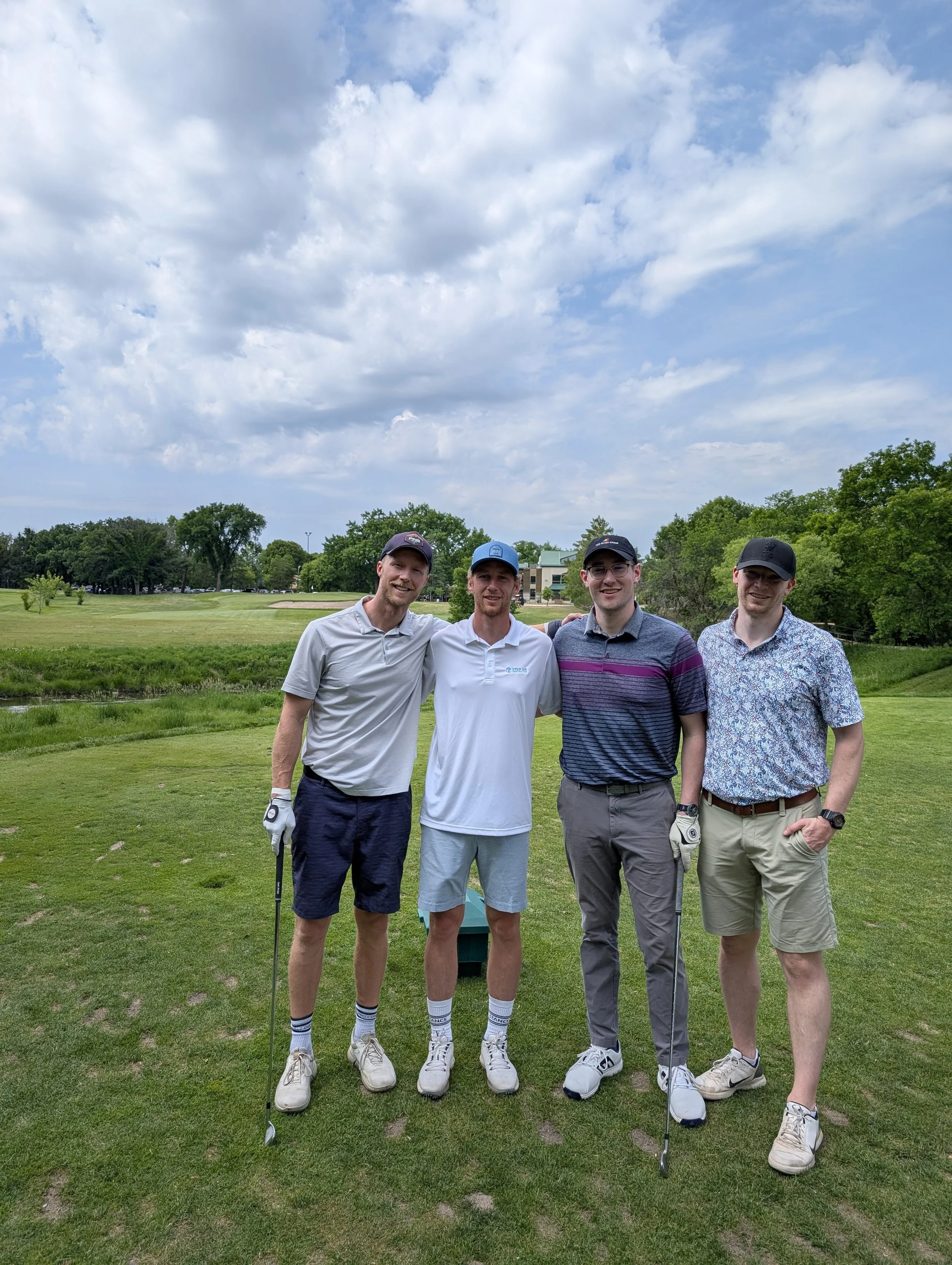 Four men standing on a golf course, smiling, with golf clubs, dressed in golf attire, and surrounded by green grass, trees, and a partly cloudy sky.
