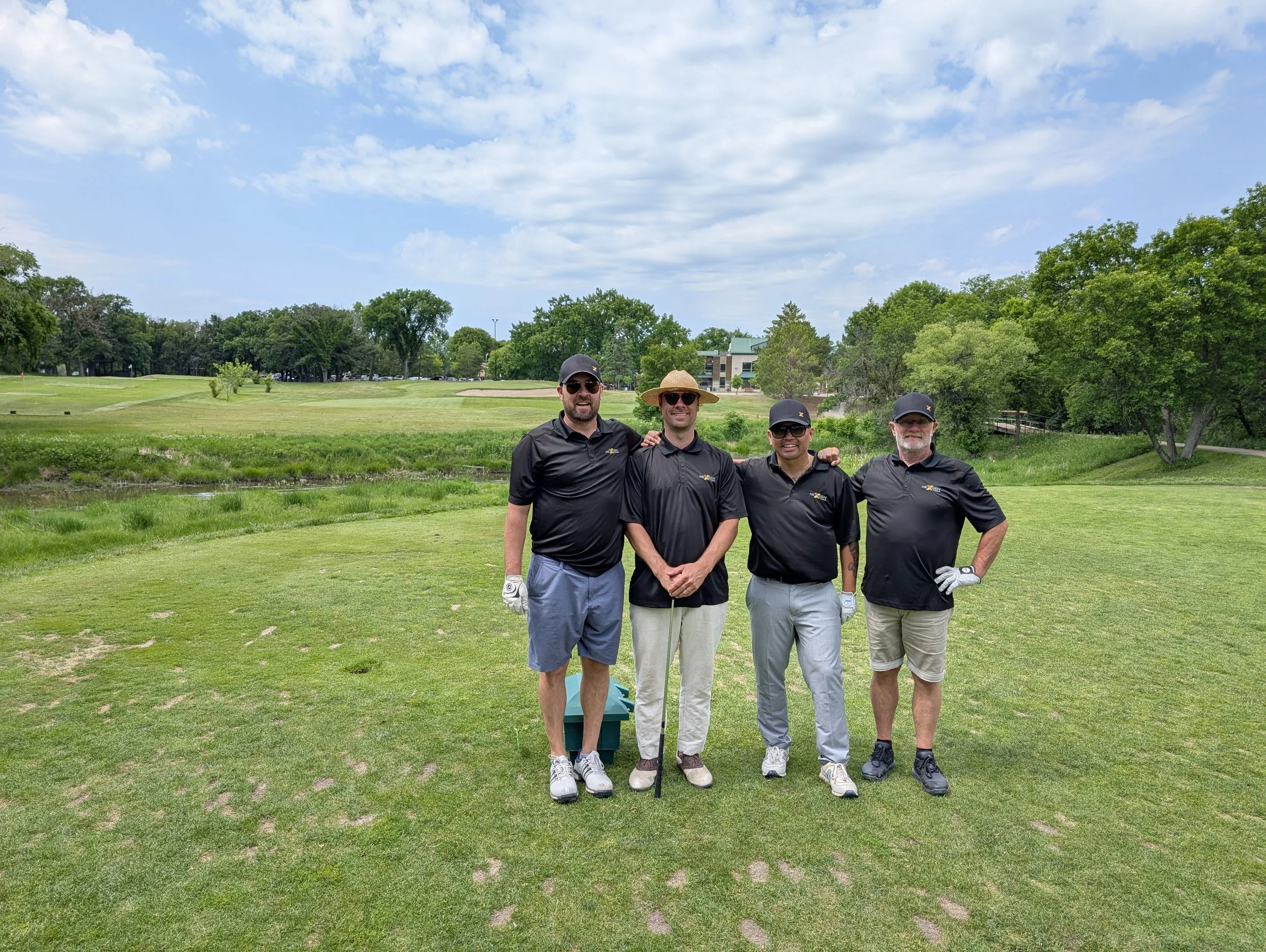 Four men dressed in black shirts and light-colored shorts or pants pose for a photo on a golf course. They are standing on the grass with golf clubs, and trees, a pond, and buildings are in the background under a partly cloudy sky.