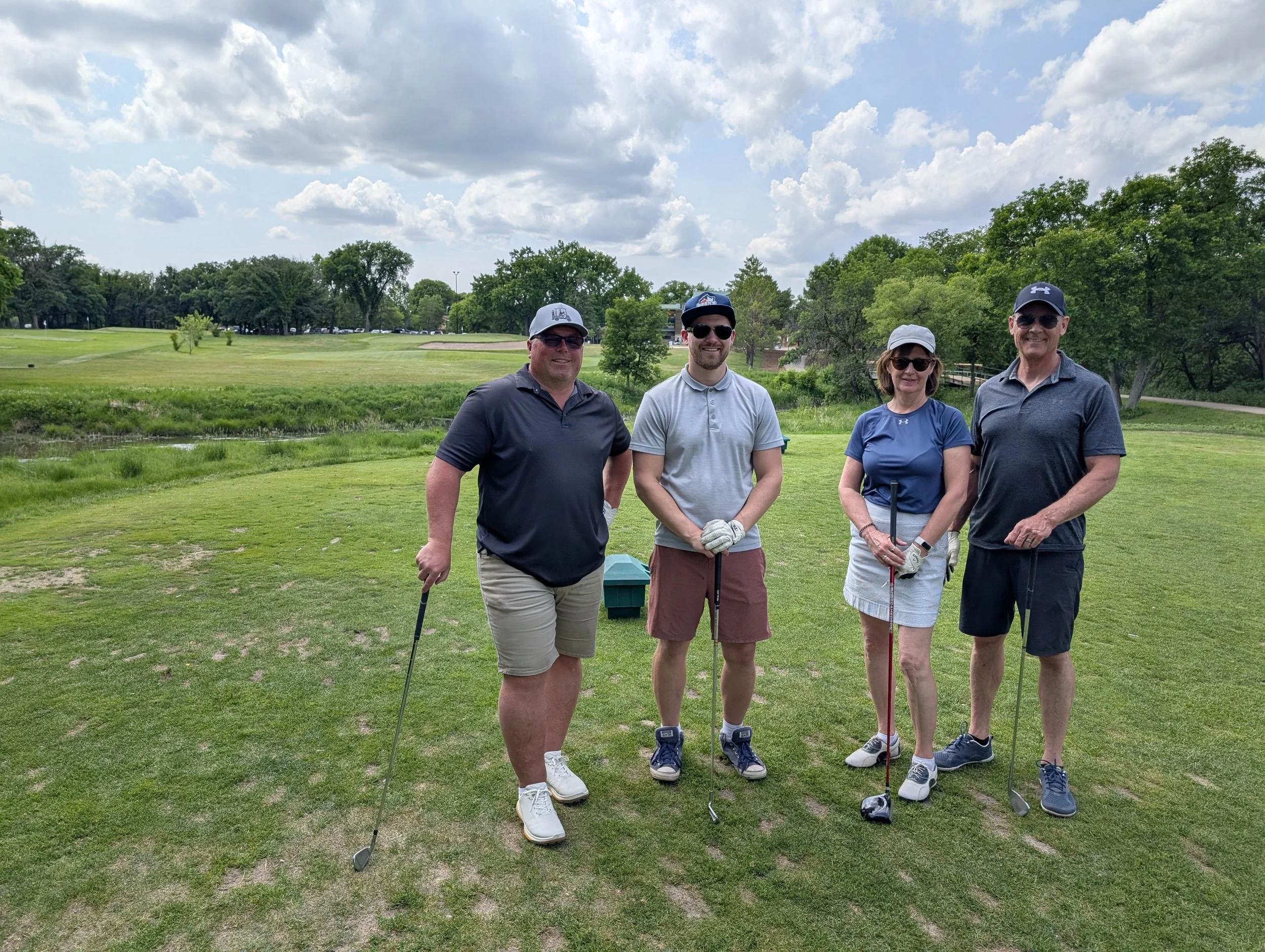 Four people standing on a golf course with golf clubs, smiling, wearing casual golf attire and sunglasses, with trees and a cloudy sky in the background.
