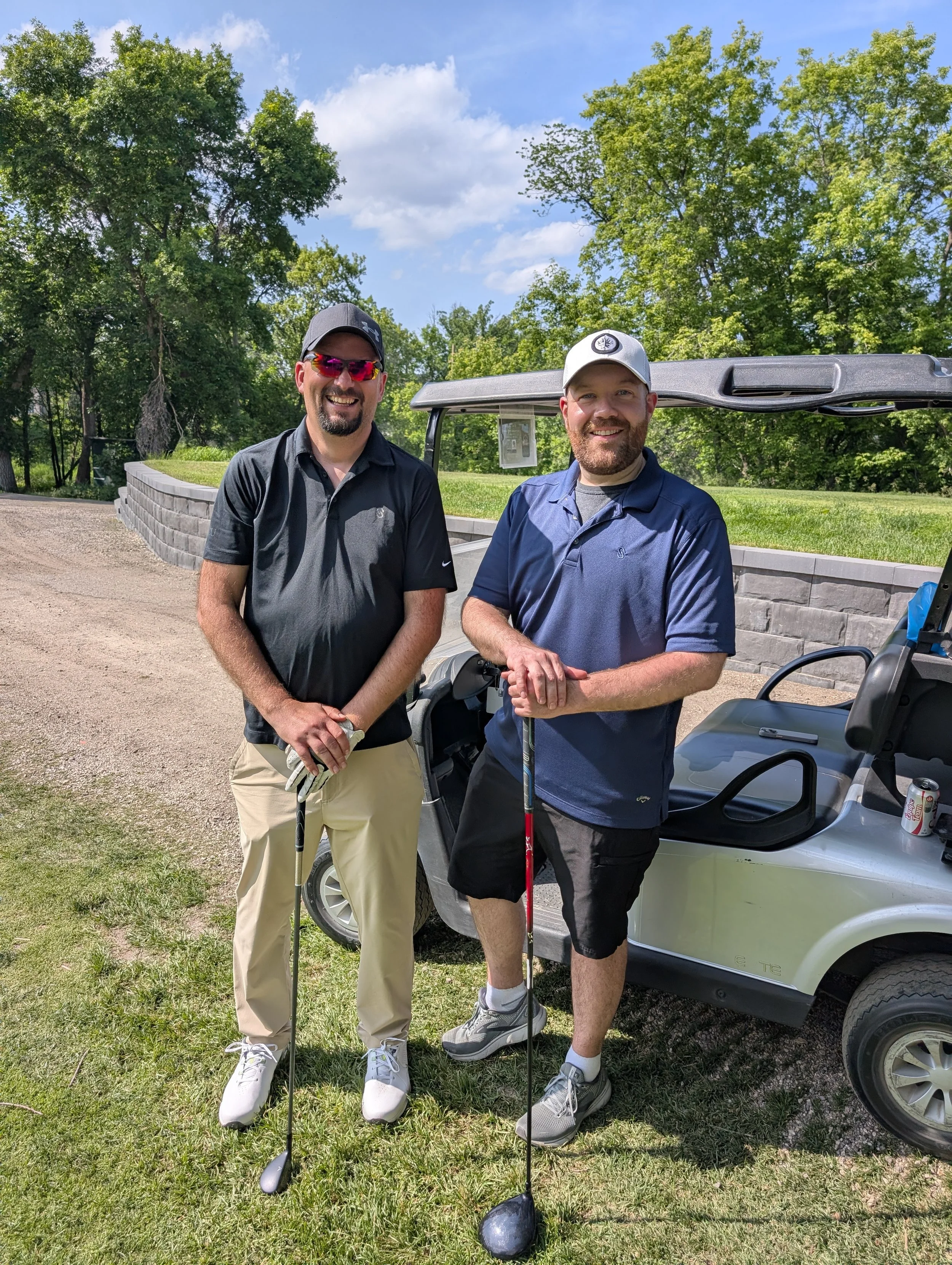 Two men smiling and posing with golf clubs on a golf course, standing next to a golf cart amid trees and a clear blue sky.