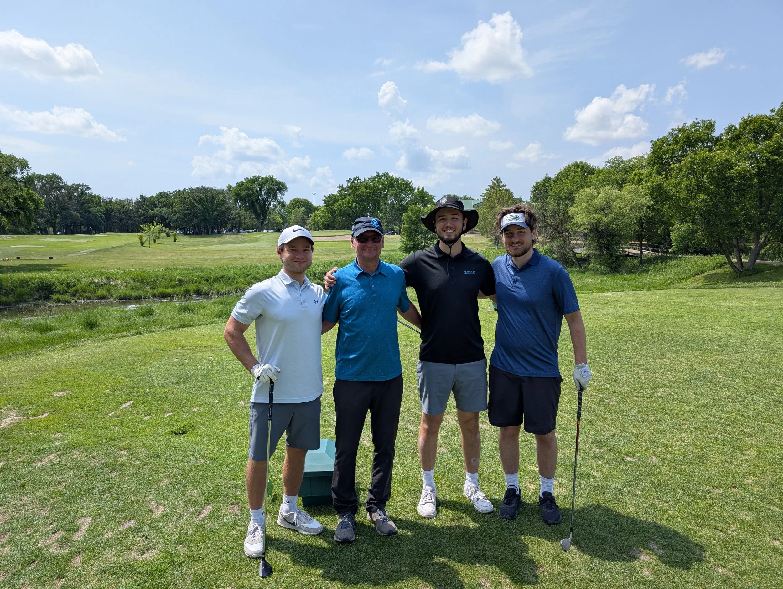 Four men in golf attire standing together on a golf course under a partly cloudy sky, smiling at the camera.