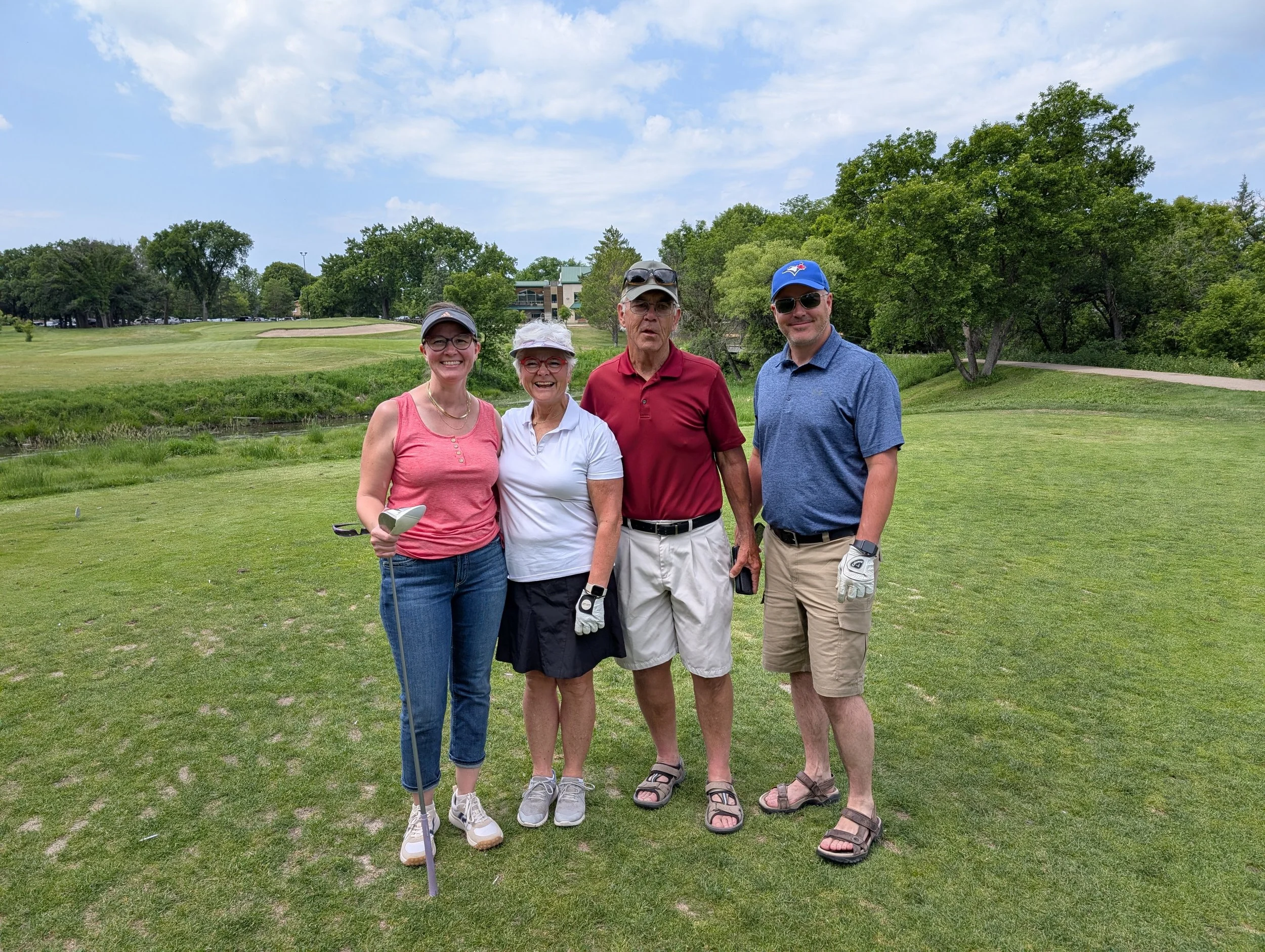 Four people standing on a golf course, posing for a photo on a bright day with clouds, trees, and a golf building in the background.
