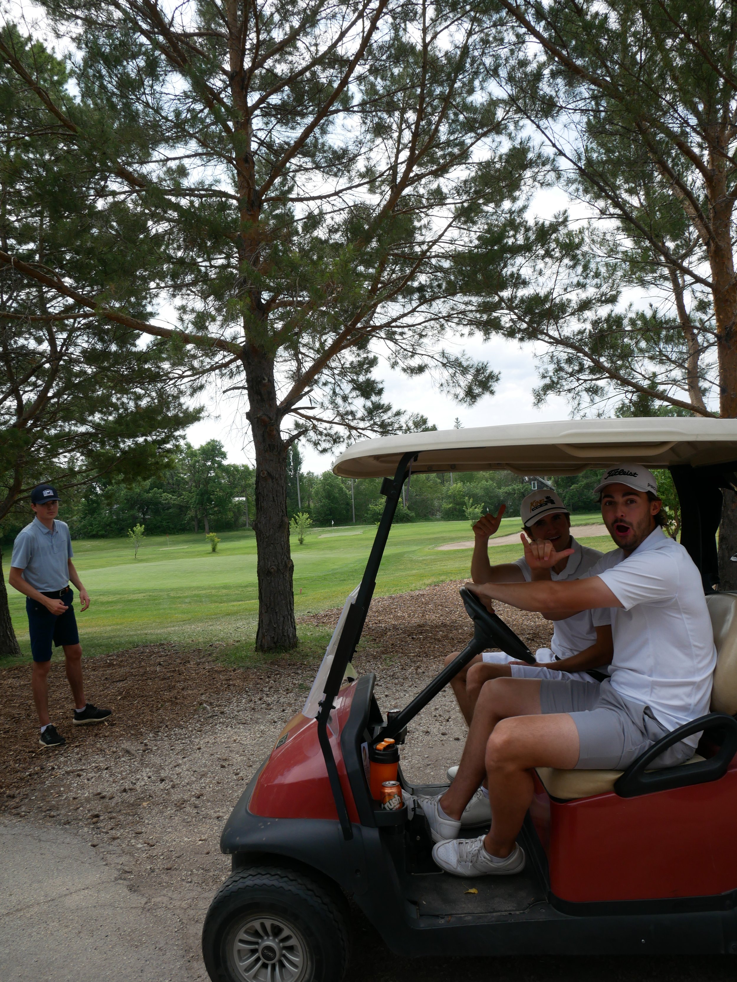 Two men sitting in a golf cart giving thumbs up, with a young boy standing nearby on a golf course surrounded by trees.
