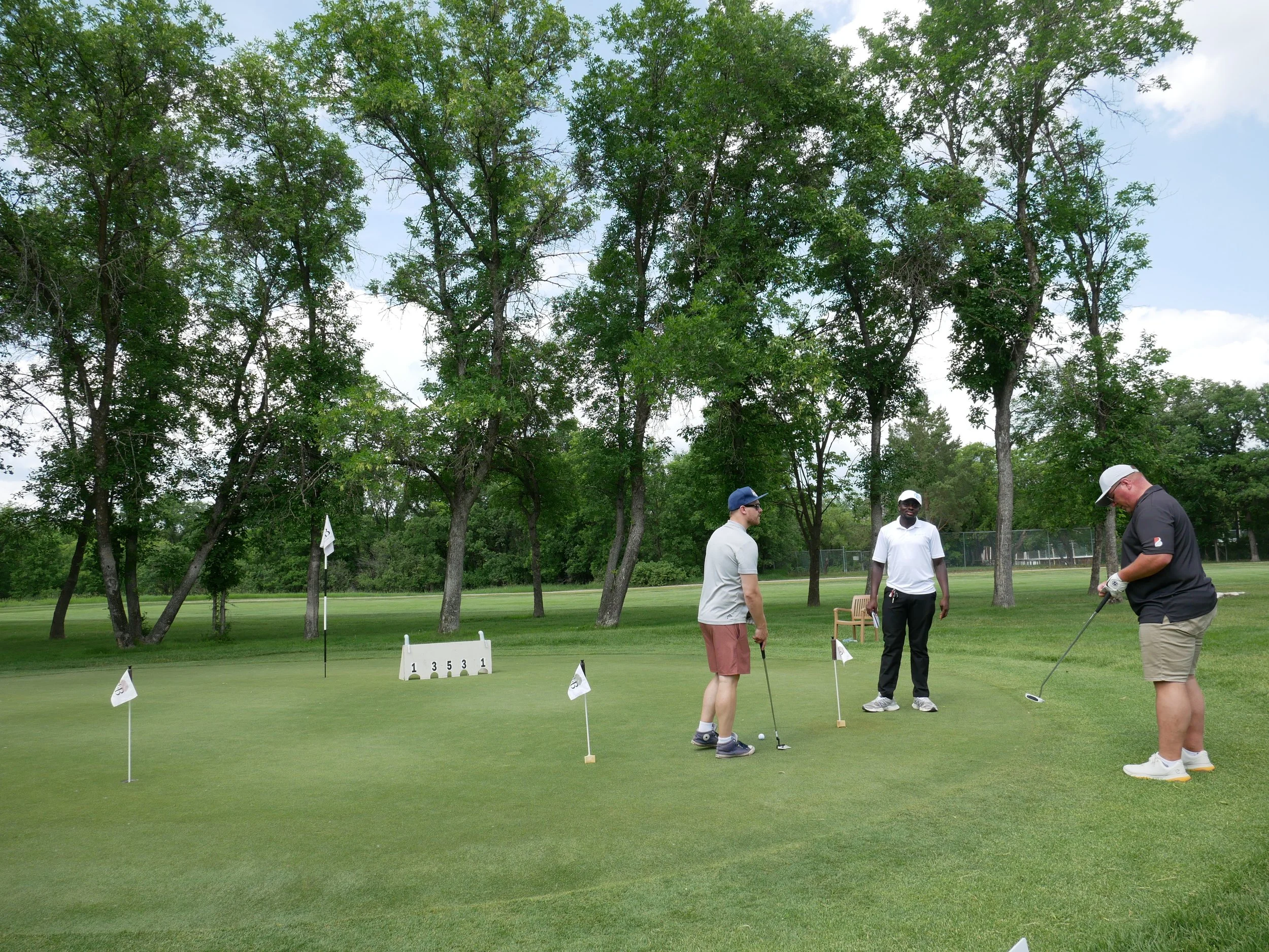 Three men playing golf on a course, with trees and a goal flag in the background.