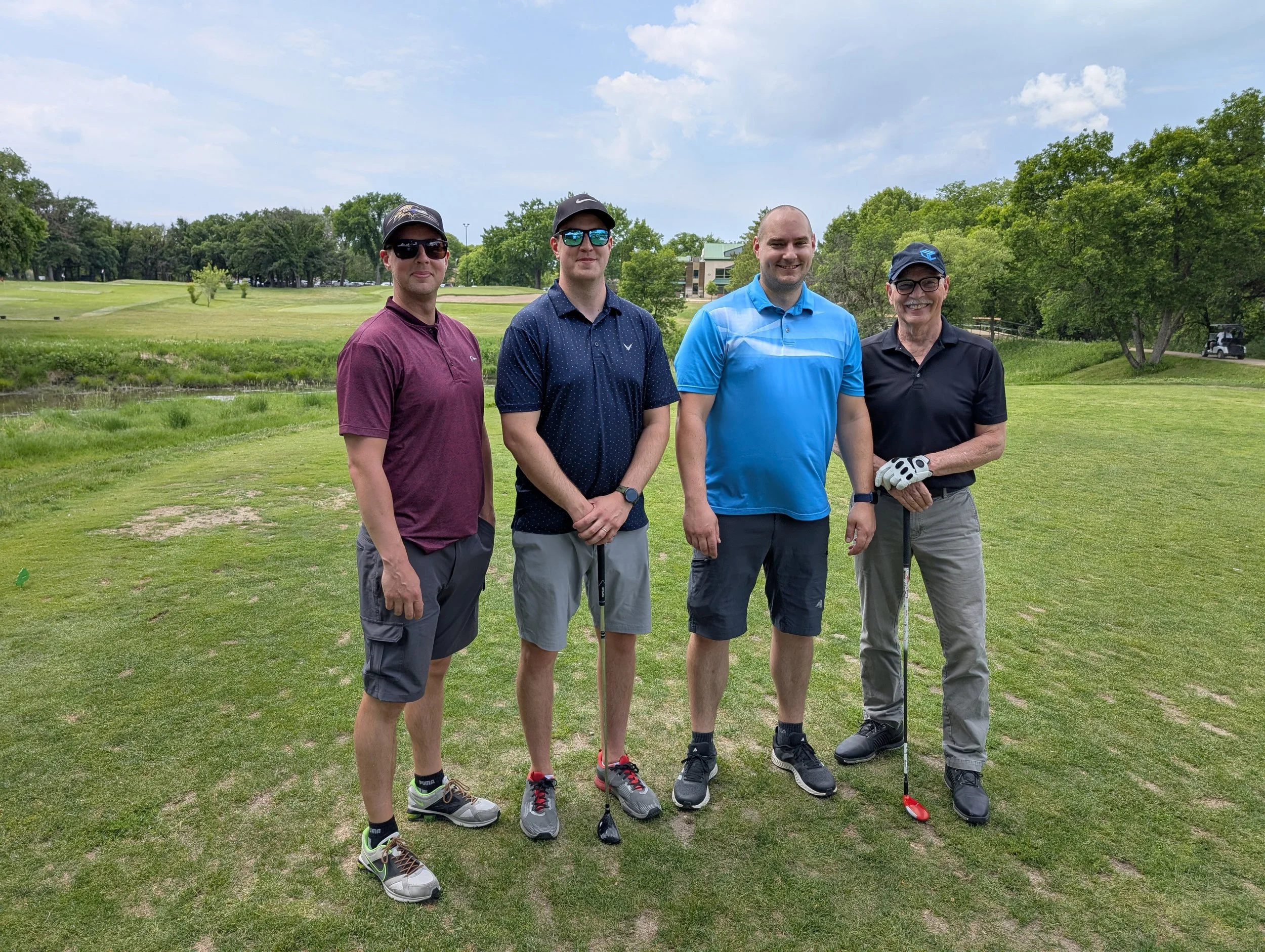 Four men standing on a golf course holding golf clubs, posing for a photo.