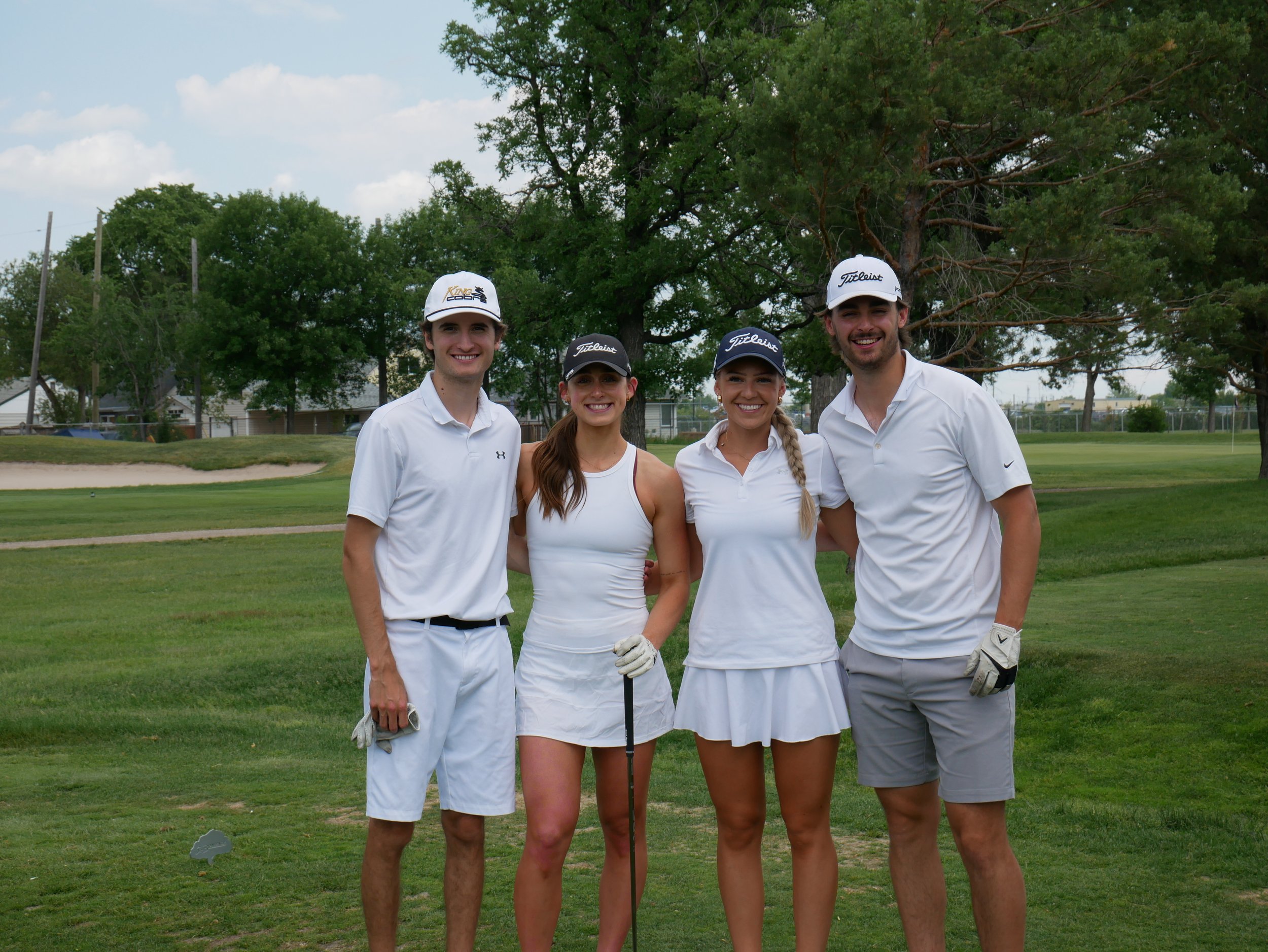 Four young adults, two men and two women, standing on a golf course wearing white golf attire and hats, smiling at the camera, with trees and golf course landscape in the background.