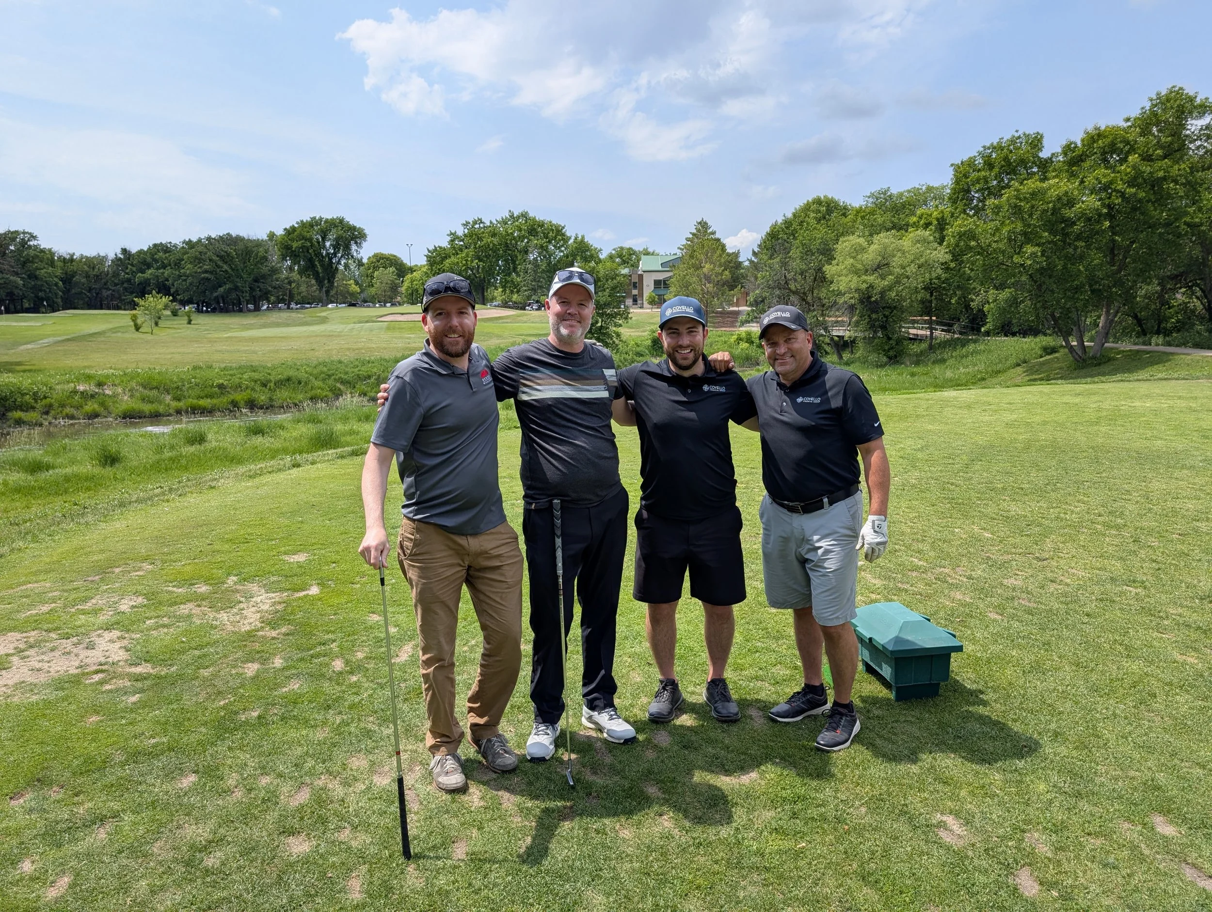 Four men standing together on a golf course, smiling, with trees and a building in the background. One man holds a golf club, and a box is on the ground nearby.