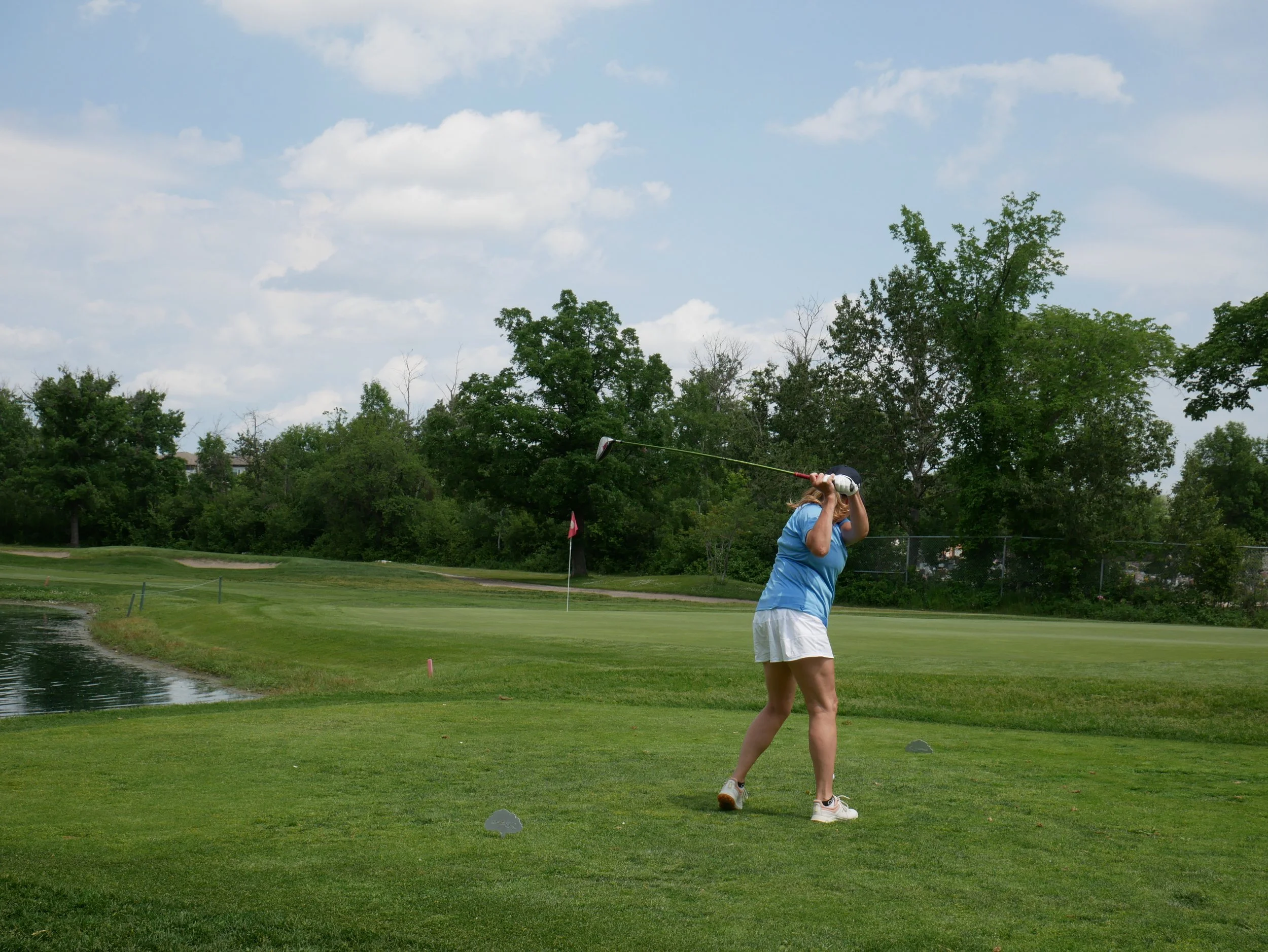 A woman in a blue shirt and white shorts swinging a golf club on a golf course. The course has green grass, a water hazard, a flag in the distance, and surrounded by trees under a partly cloudy sky.