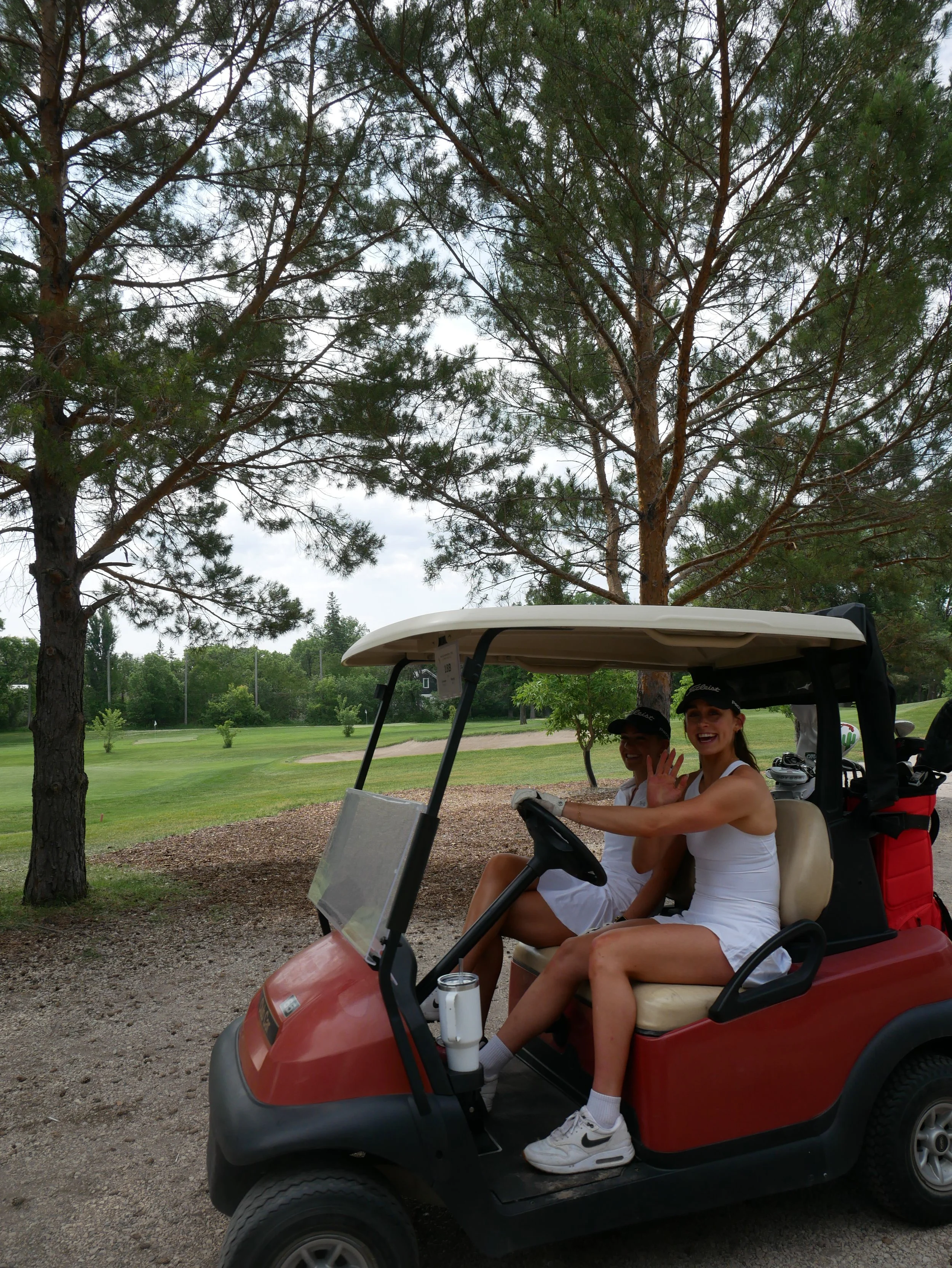 Two women in white athletic outfits and caps sitting in a red golf cart on a golf course, smiling and waving at the camera, with trees and a green field in the background.