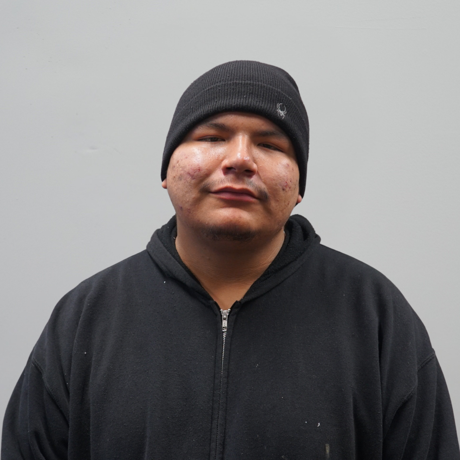 A young man with glasses and black spiky hair wearing a black hoodie with the word 'WORK' and a blue 3D cube logo in the middle, standing in front of closed white blinds.