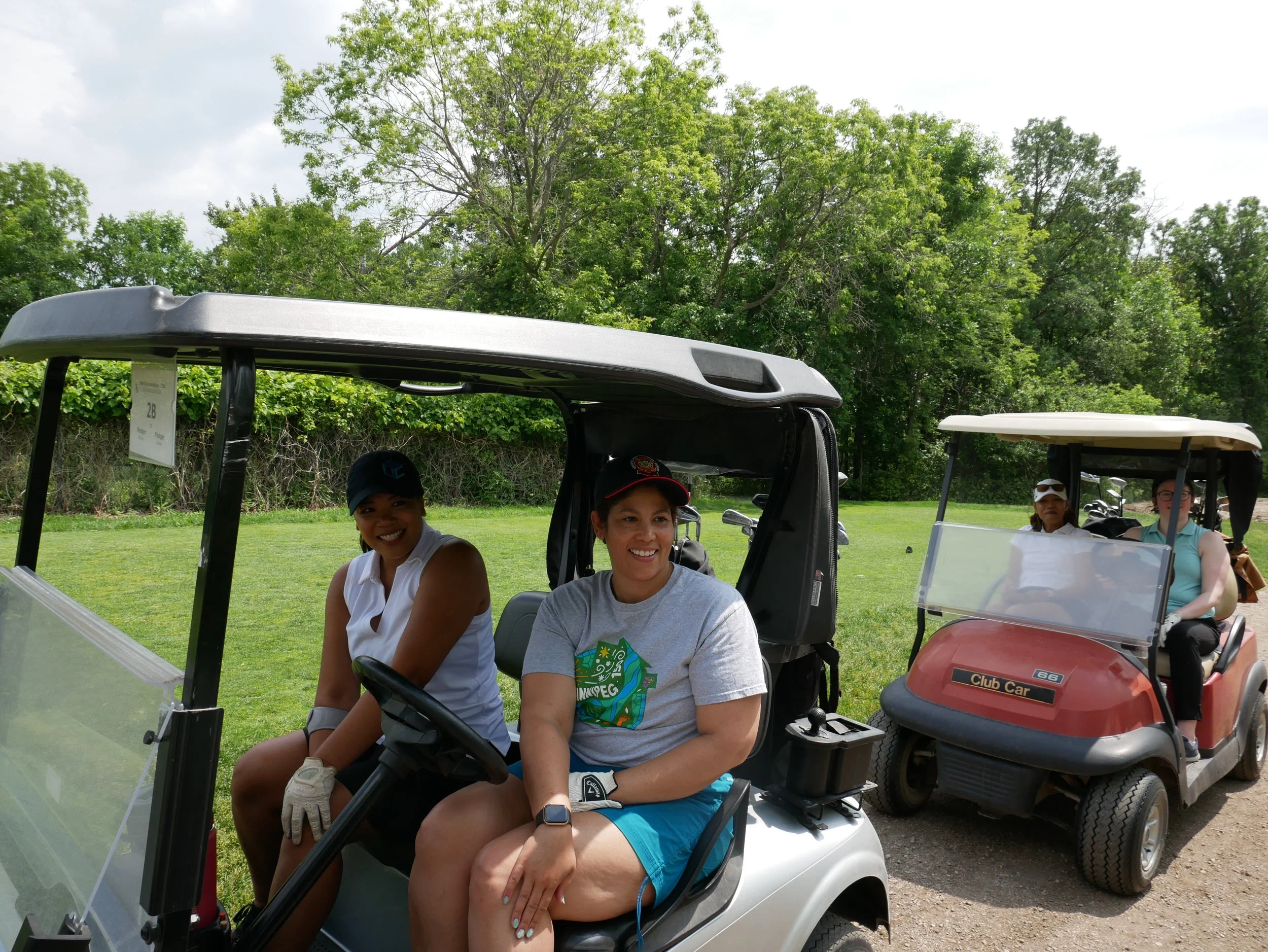 Women riding in two golf carts on a golf course with green grass and trees in the background.