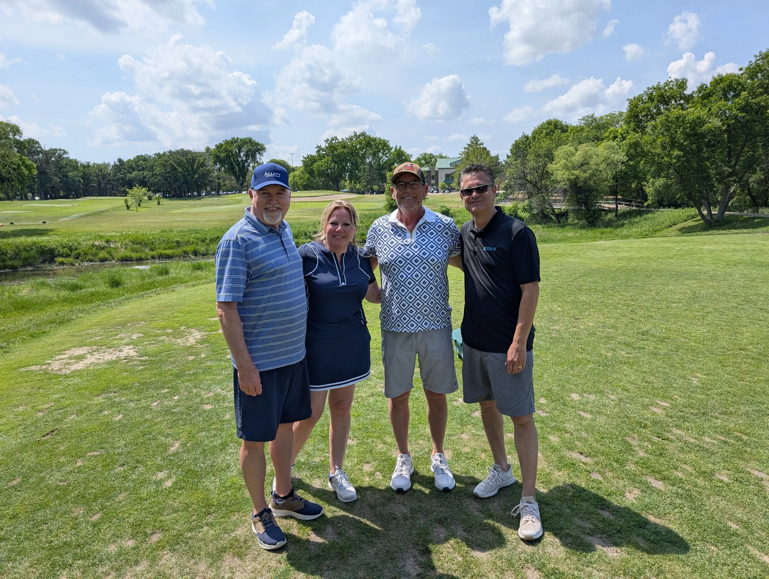 Four people standing on a golf course on a sunny day, smiling for the camera, with trees and a building in the background.
