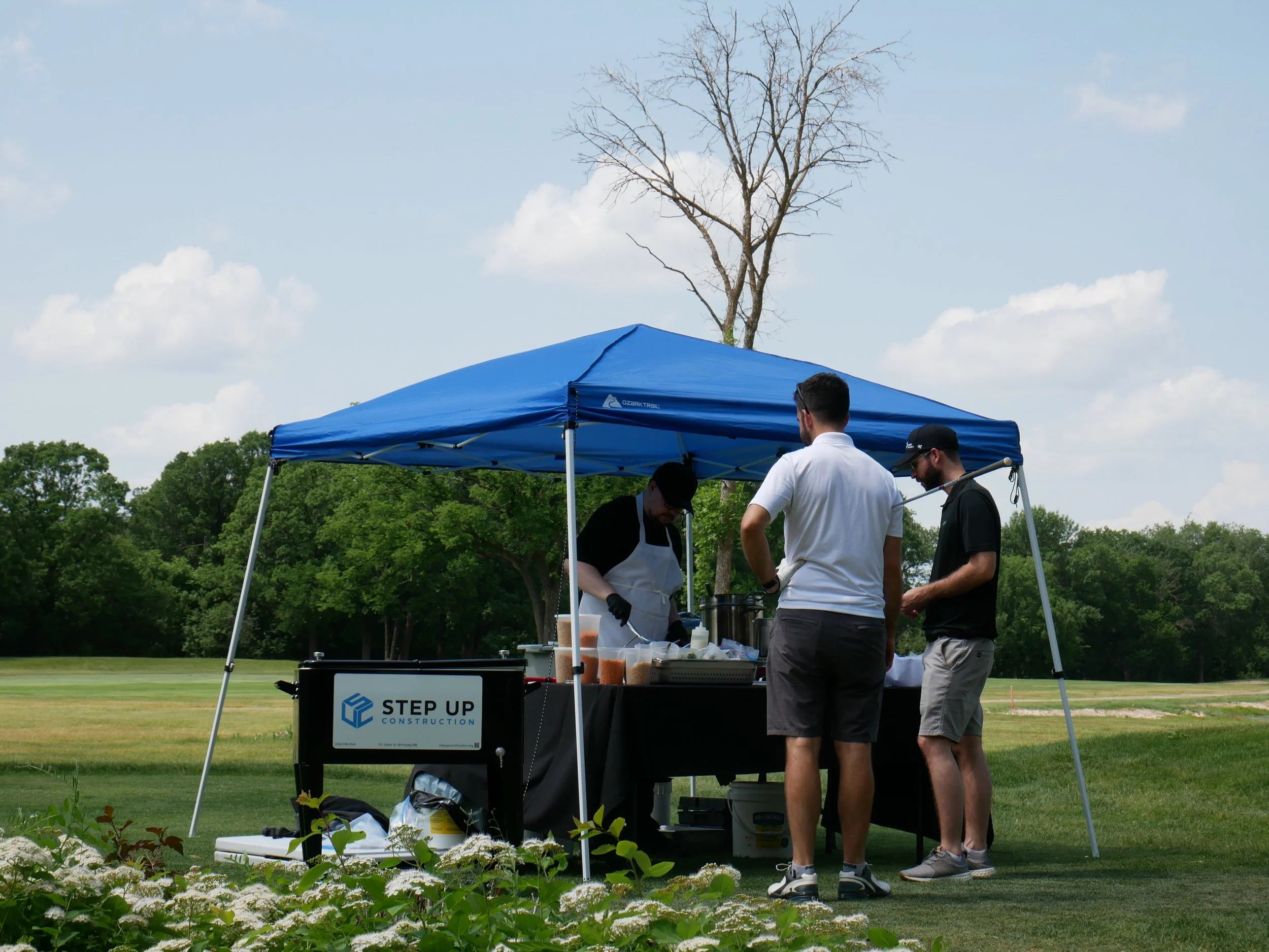 Three men at a food stand under a blue canopy in a grassy outdoor setting with trees and a partly cloudy sky in the background.