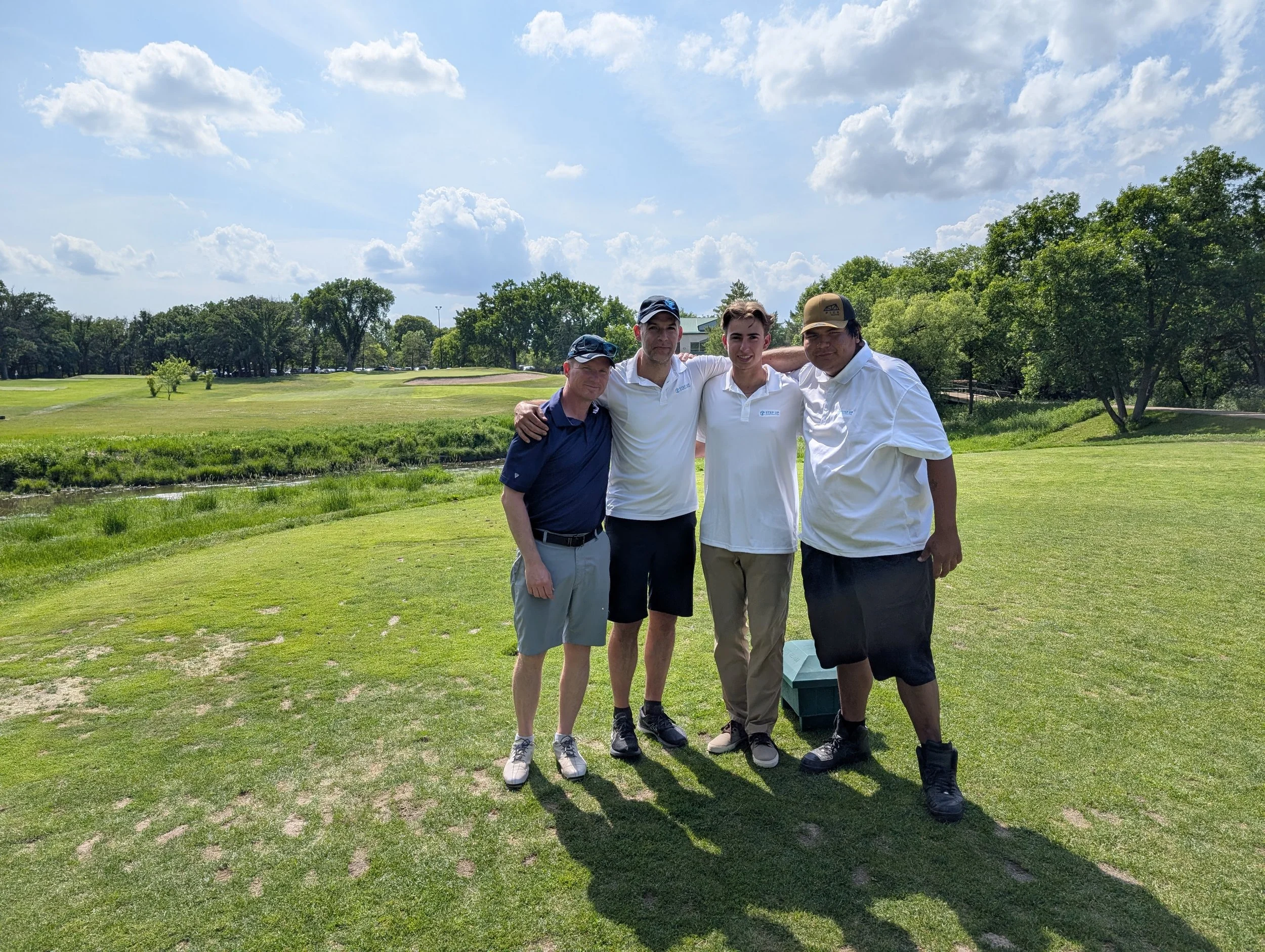 Four men standing together on a golf course, with lush green grass, trees, and a partly cloudy sky in the background. All are wearing polo shirts and shorts.