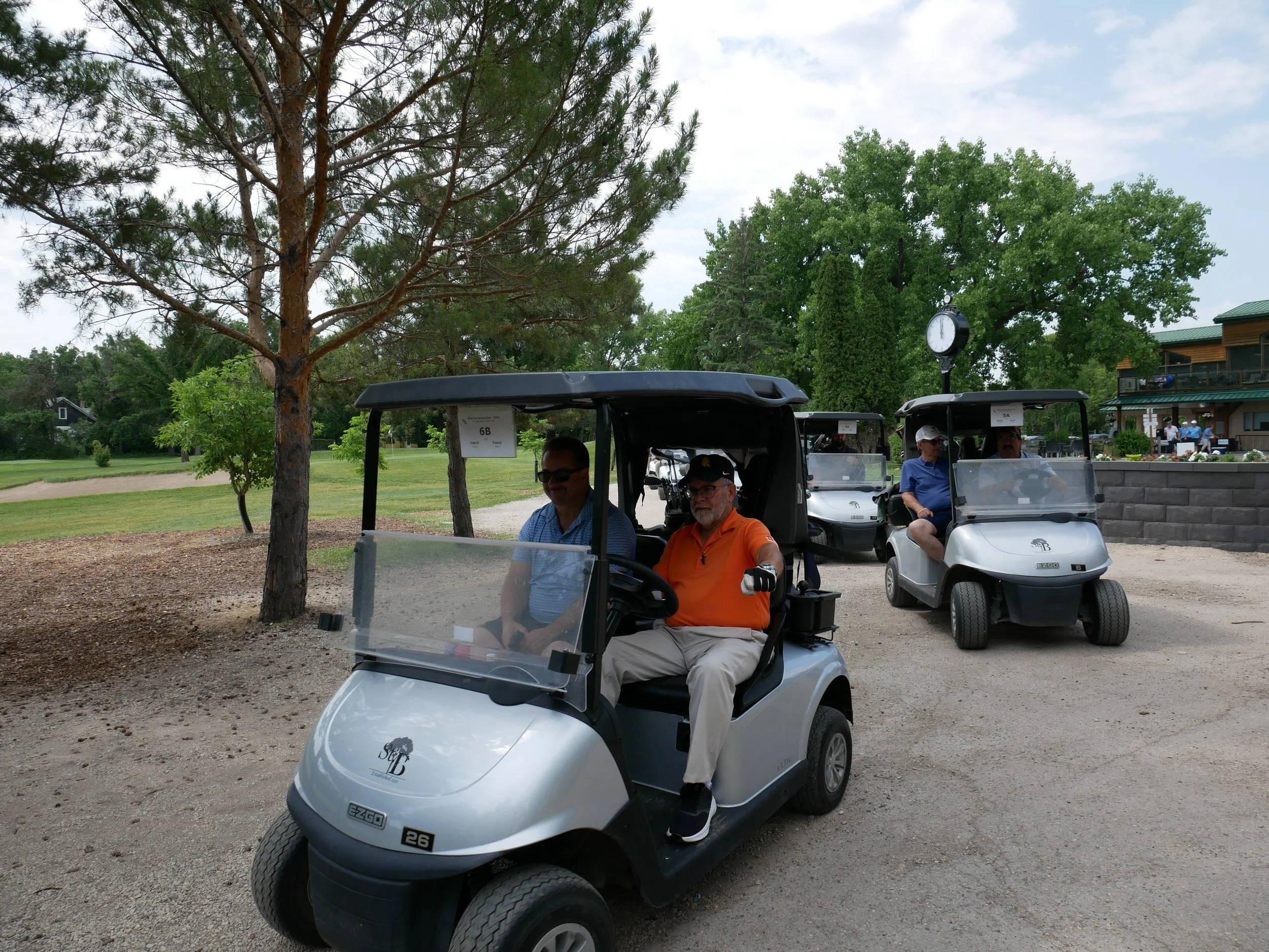 Four men riding in golf carts on a golf course with trees, grass, and a building in the background.