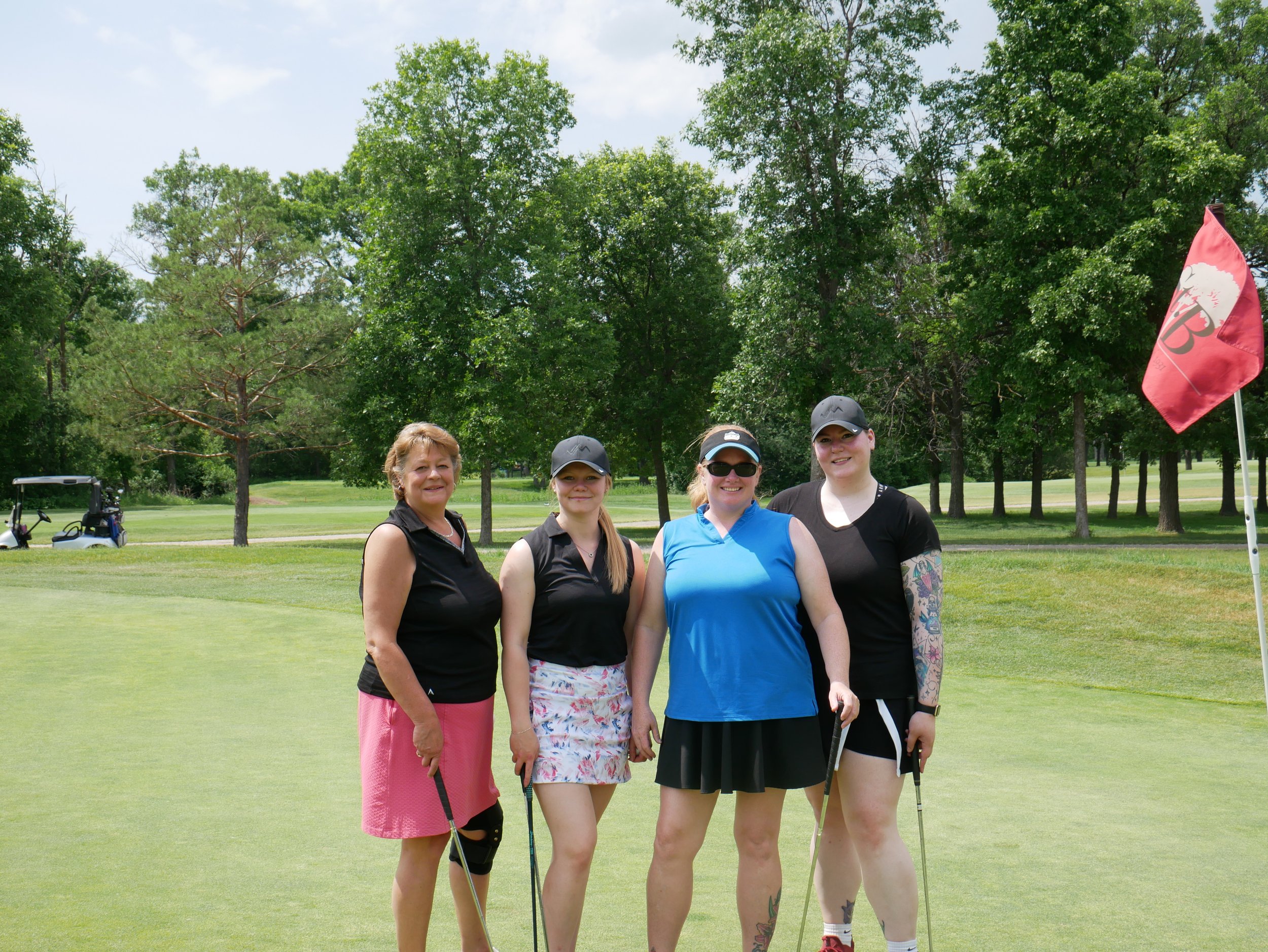 Four women standing together on a golf course near a flag, holding golf clubs and smiling, with green trees and a golf cart in the background.