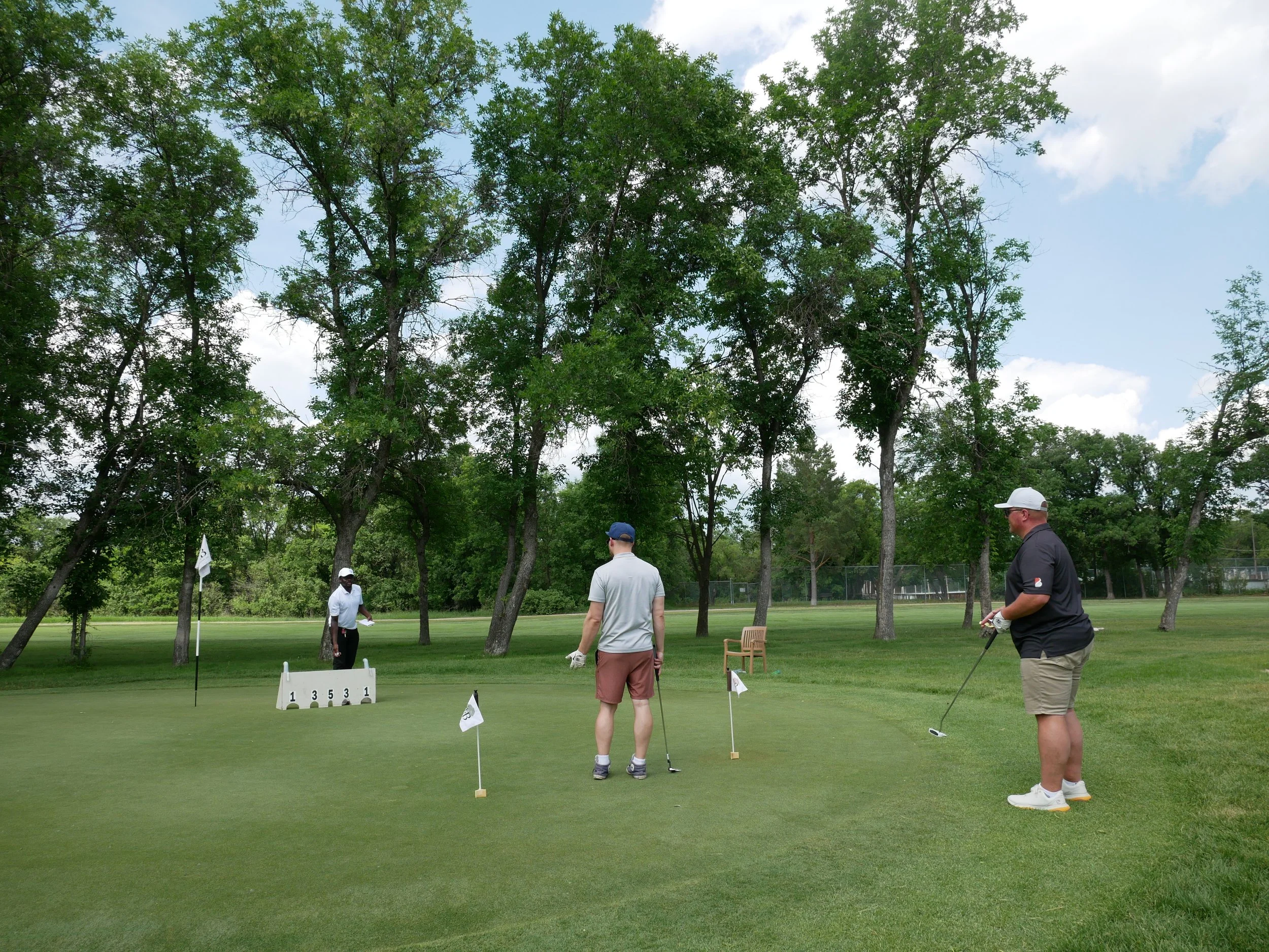 Three men on a golf course preparing to putt, with a sign for a golf game scoring or rules in front of them and trees in the background.