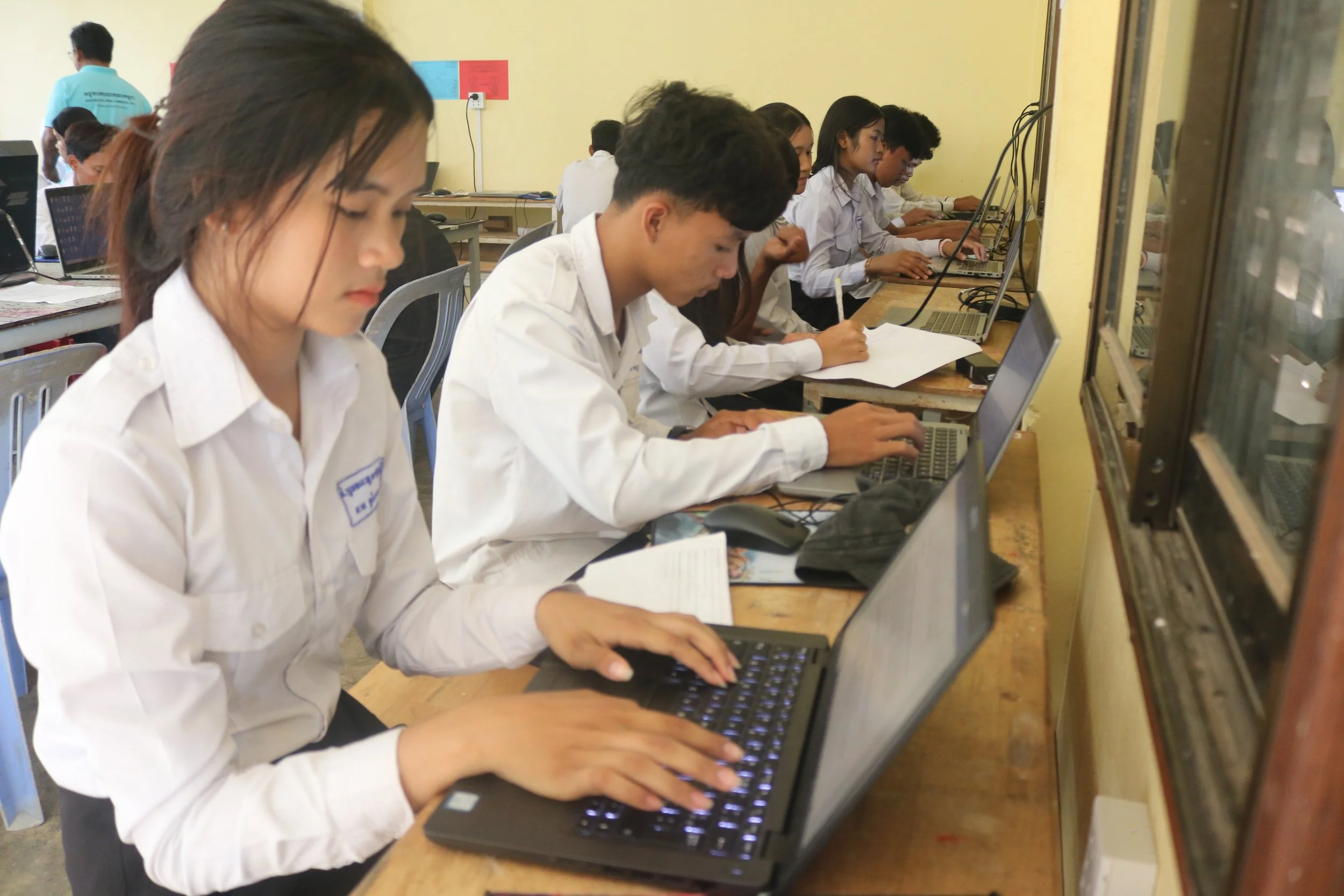 A classroom with students working on desktop computers, some students are paired and focused on their screens, while a teacher or supervisor stands nearby. The room has yellow walls, windows with bars, and posters on the wall.