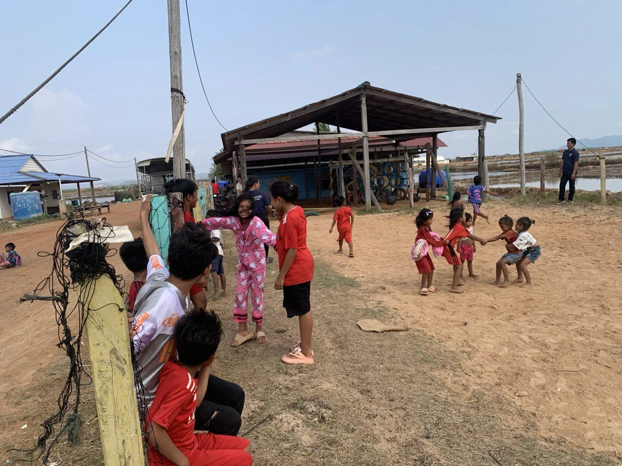 Children playing tug-of-war and running in a dirt playground near a wooden structure, with adults sitting and standing nearby, in a rural area with water paddies and simple houses.