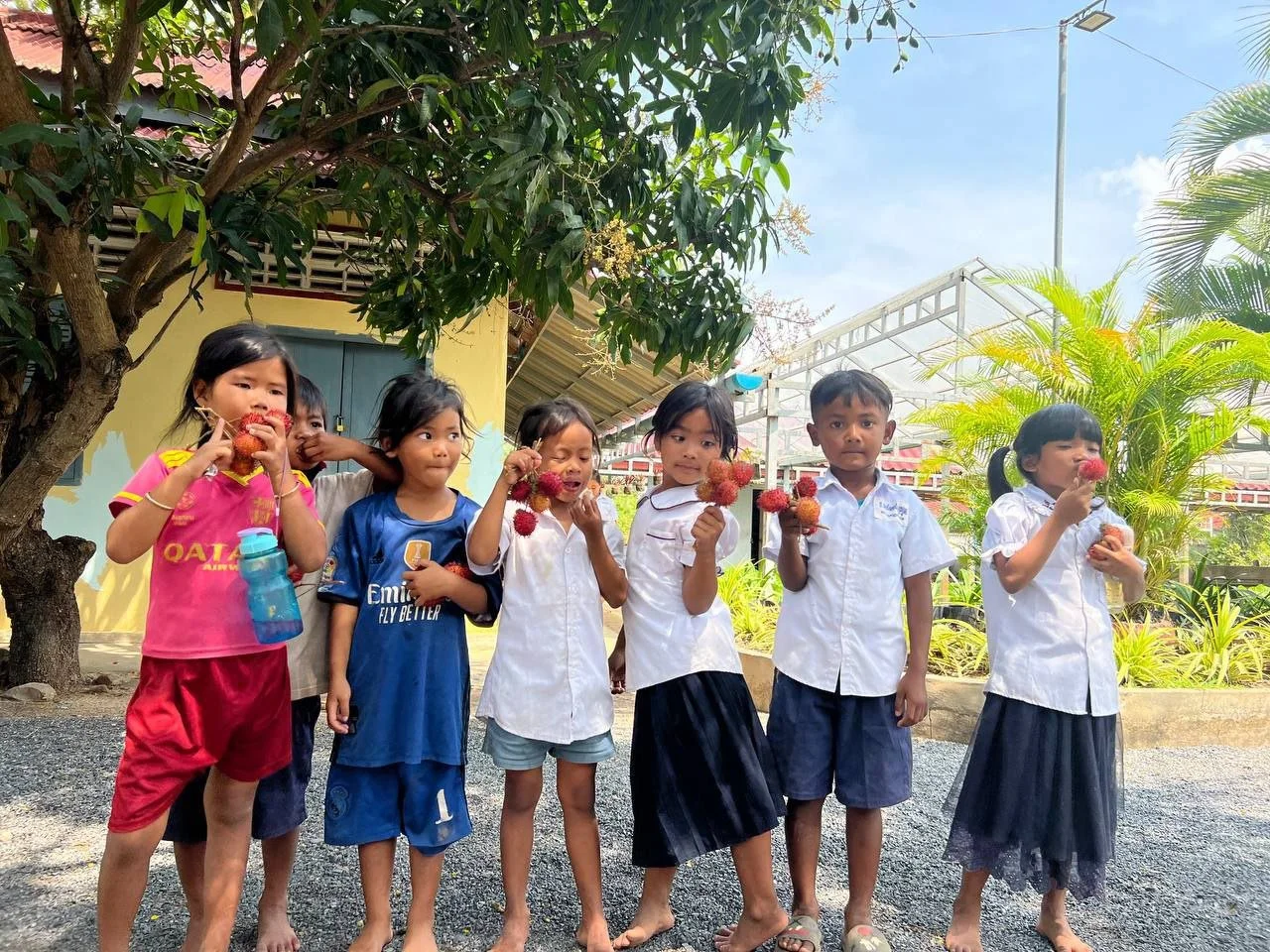 Seven children standing outdoors, holding and eating red and orange flower-like objects, with trees and a building with a yellow wall in the background.
