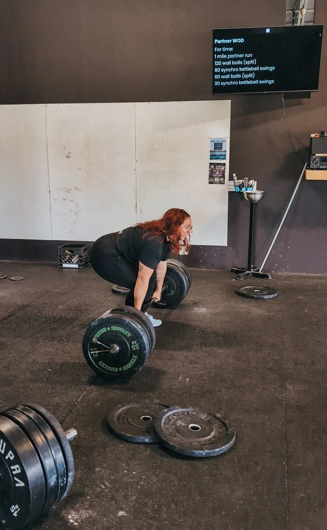 A woman with red hair lifting a heavy barbell in a gym, with weight plates on the ground nearby and a digital workout timer on a screen on the wall.