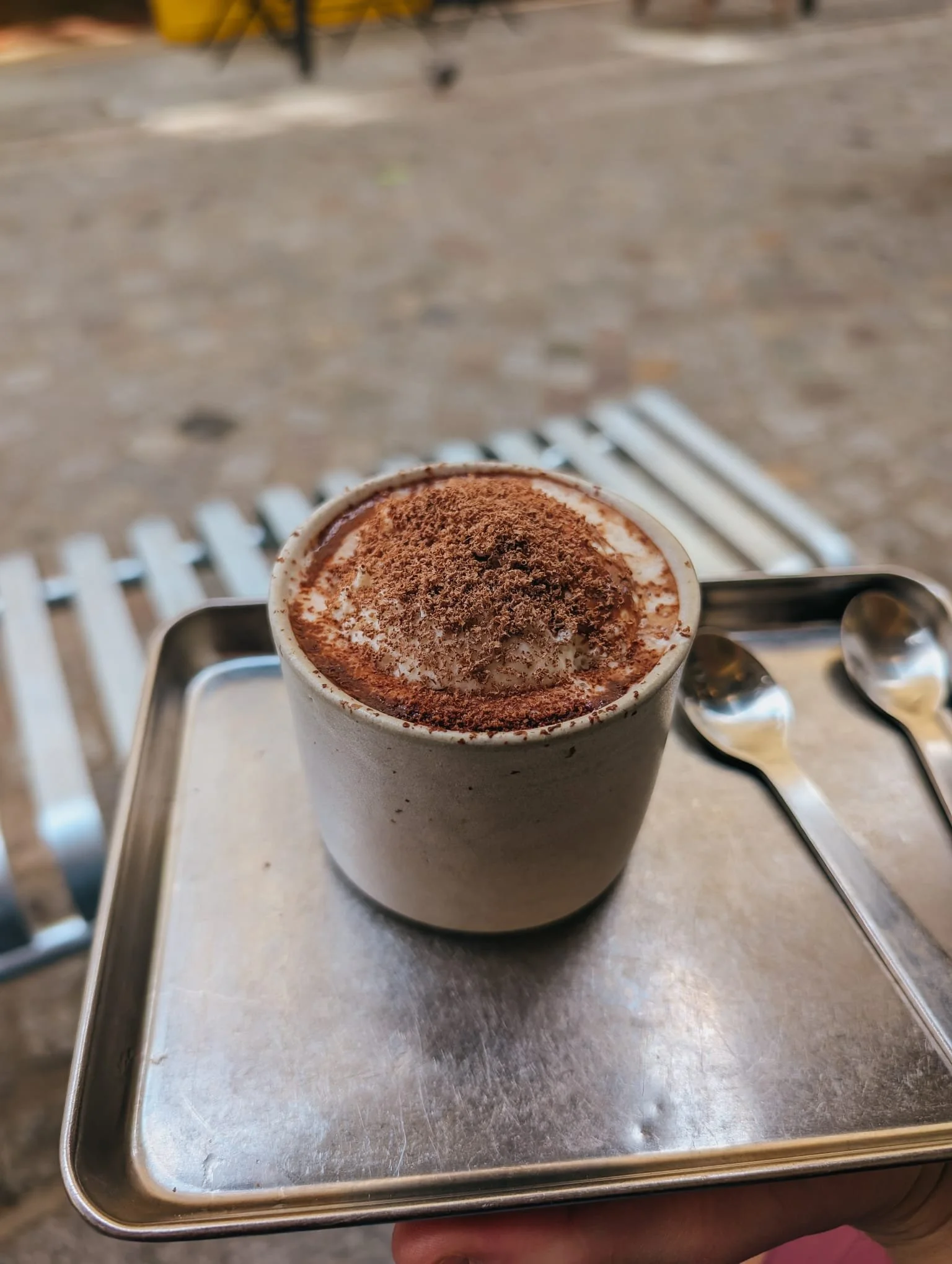 A hot beverage, possibly coffee or hot chocolate, topped with whipped cream and sprinkled with cocoa powder or chocolate shavings, served in a white ceramic mug. The mug is placed on a metal tray with two small spoons next to it.