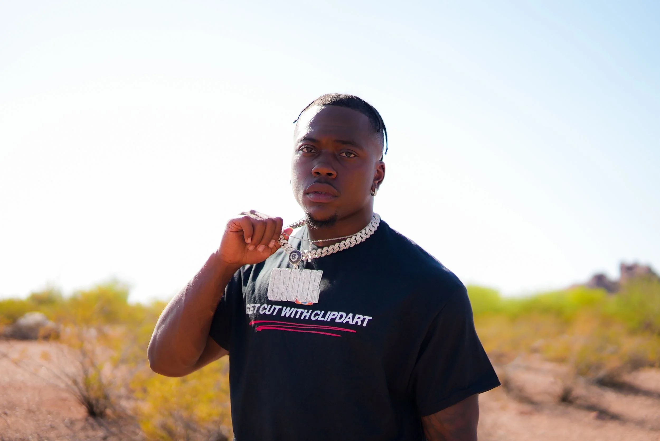 A young man with a serious expression outdoors in a desert-like area during daytime. He's wearing a black t-shirt with writing, a chunky chain necklace, and earrings, and is holding up his right hand near his shoulder.