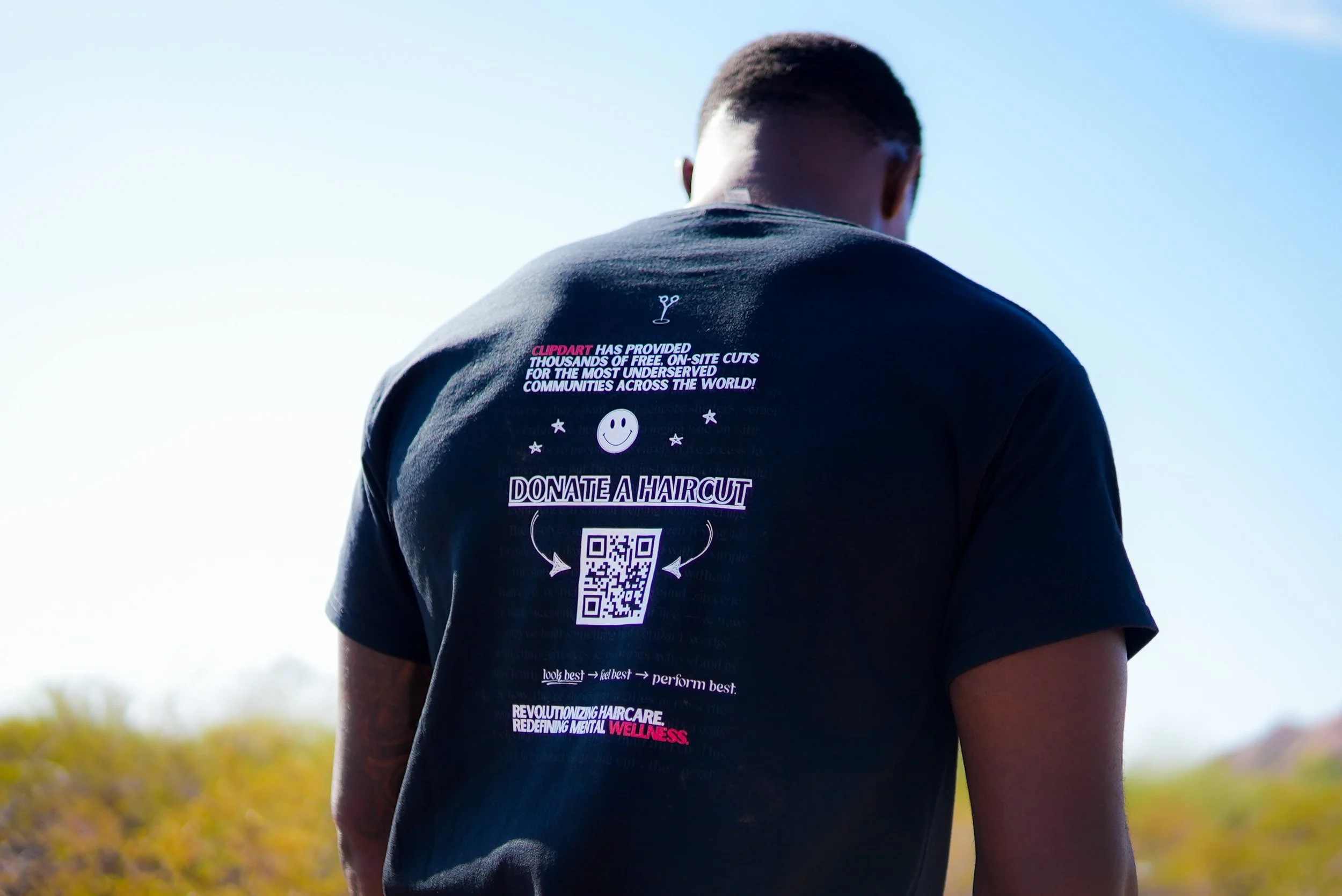 Back of a person's T-shirt with text promoting a haircut donation campaign, featuring a QR code and colorful design elements.