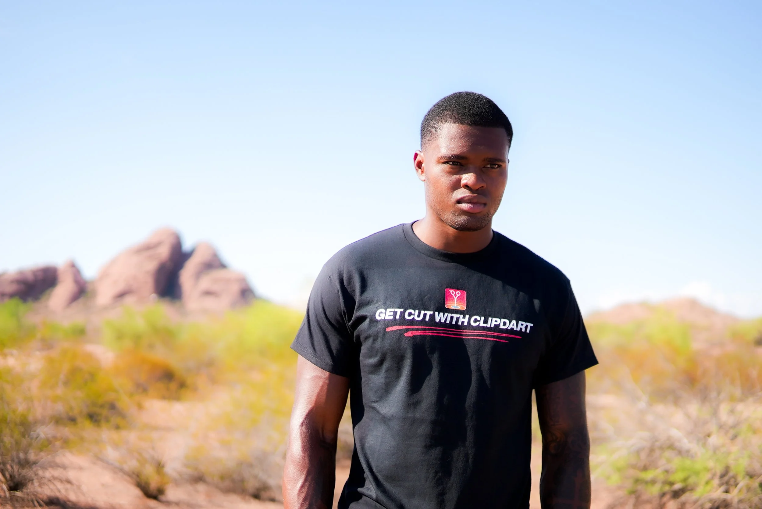 A young man outdoors in a desert landscape, wearing a black T-shirt with the text 'GET CUT WITH CLIPDART' and a pair of scissors above the text.