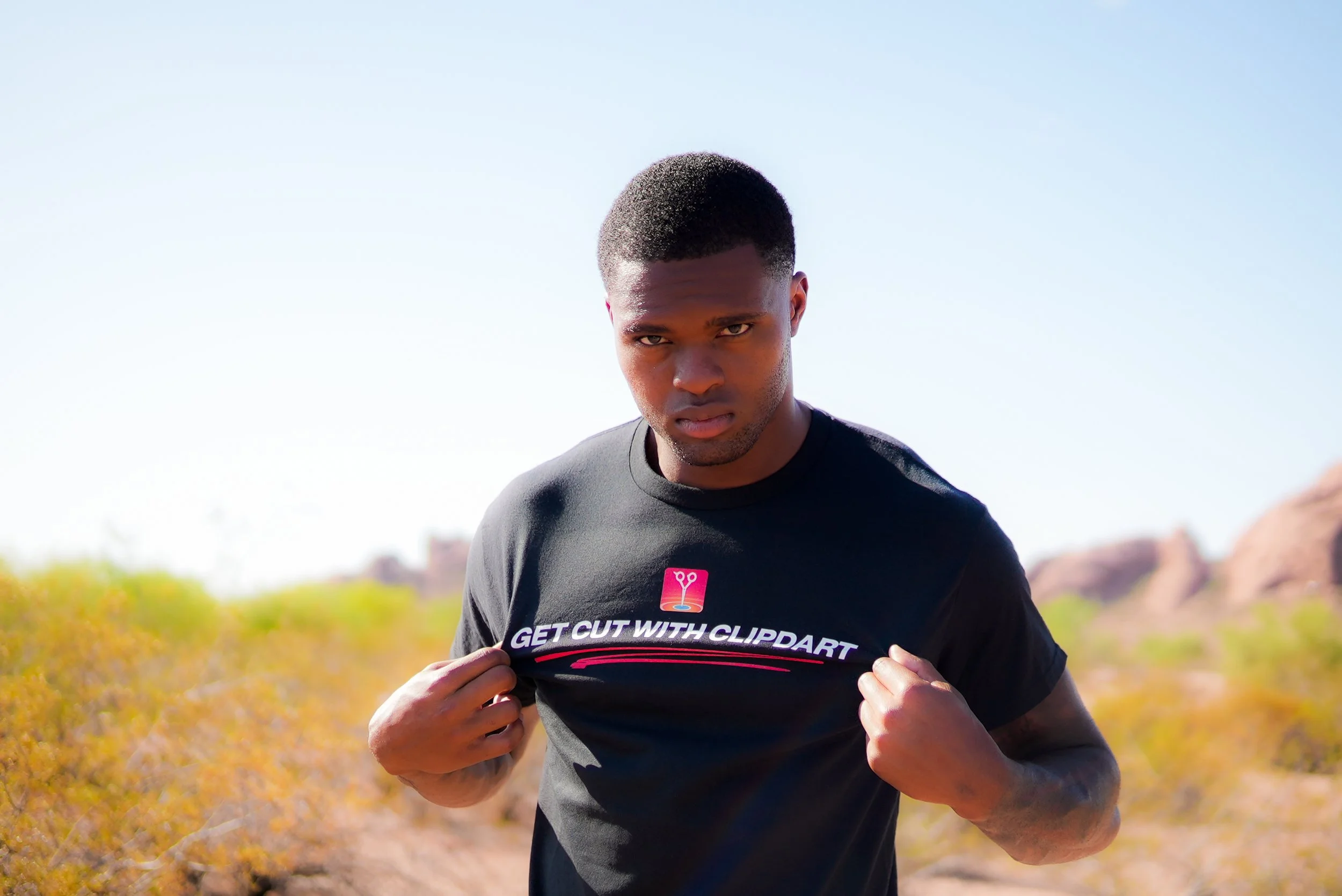 Young man outdoors in desert landscape with rocks and shrubs, wearing a black T-shirt that says 'Get Cut with Clippart' and staring into camera.