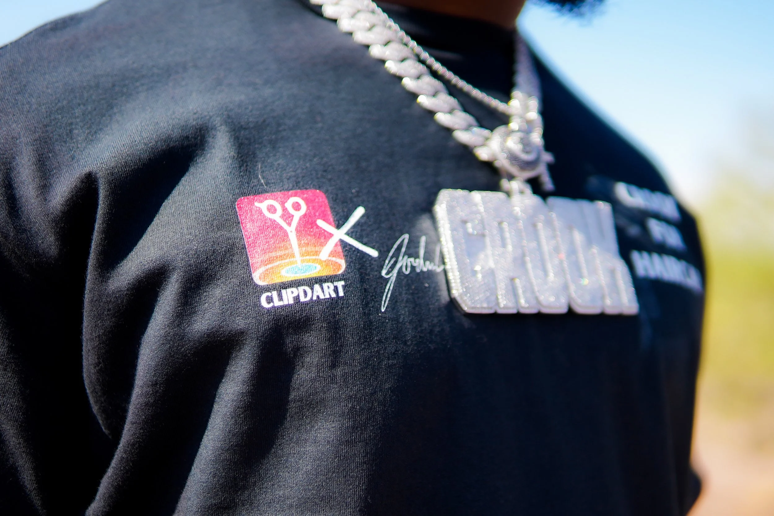 Close-up of a person wearing a black shirt with a large silver chain with a rectangular pendant and a patch that reads 'CLIPART' with a logo of scissors cutting a line.