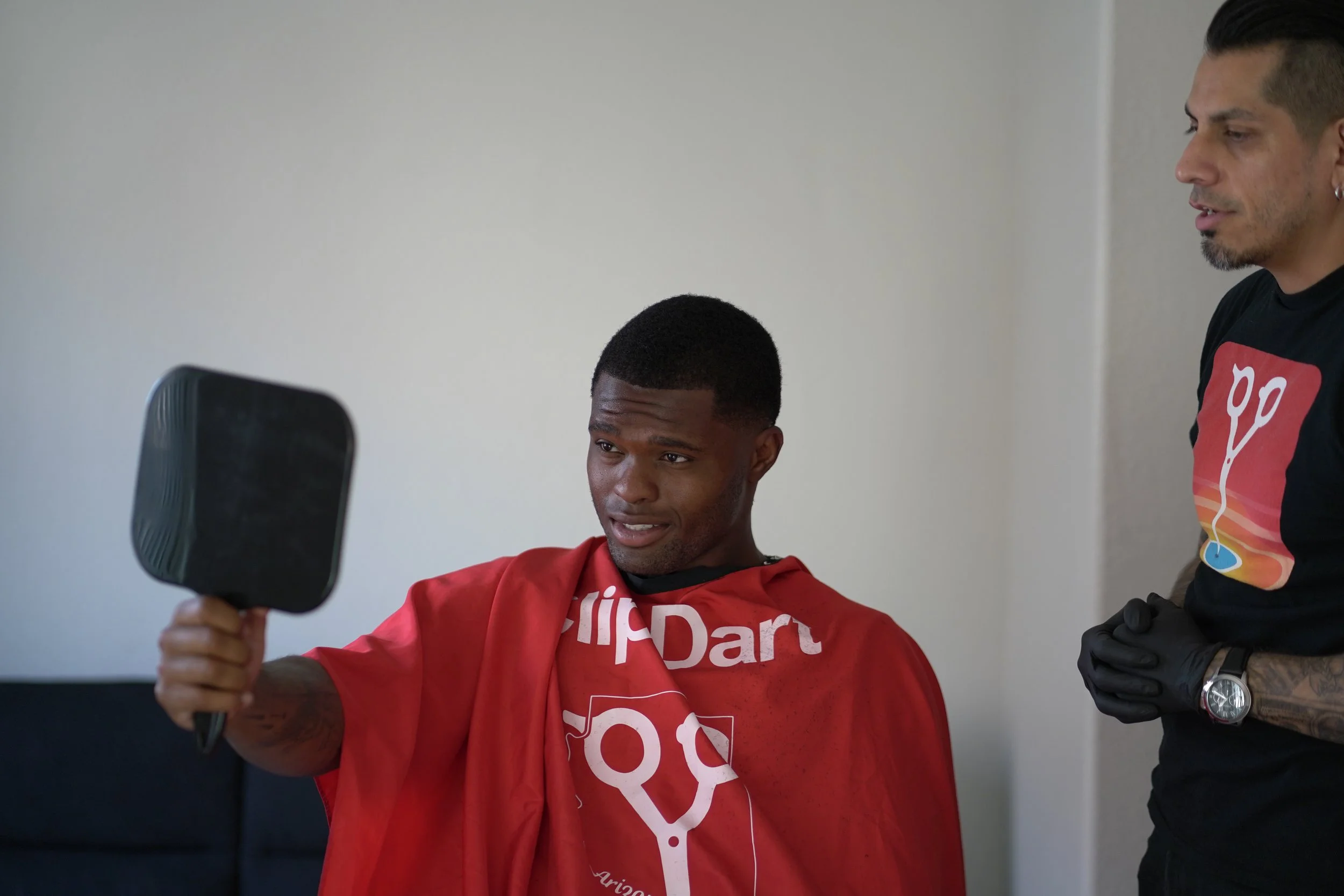 A man sitting in a barber's chair, wearing a red cape with white graphics, looking at a mirror held by another person.