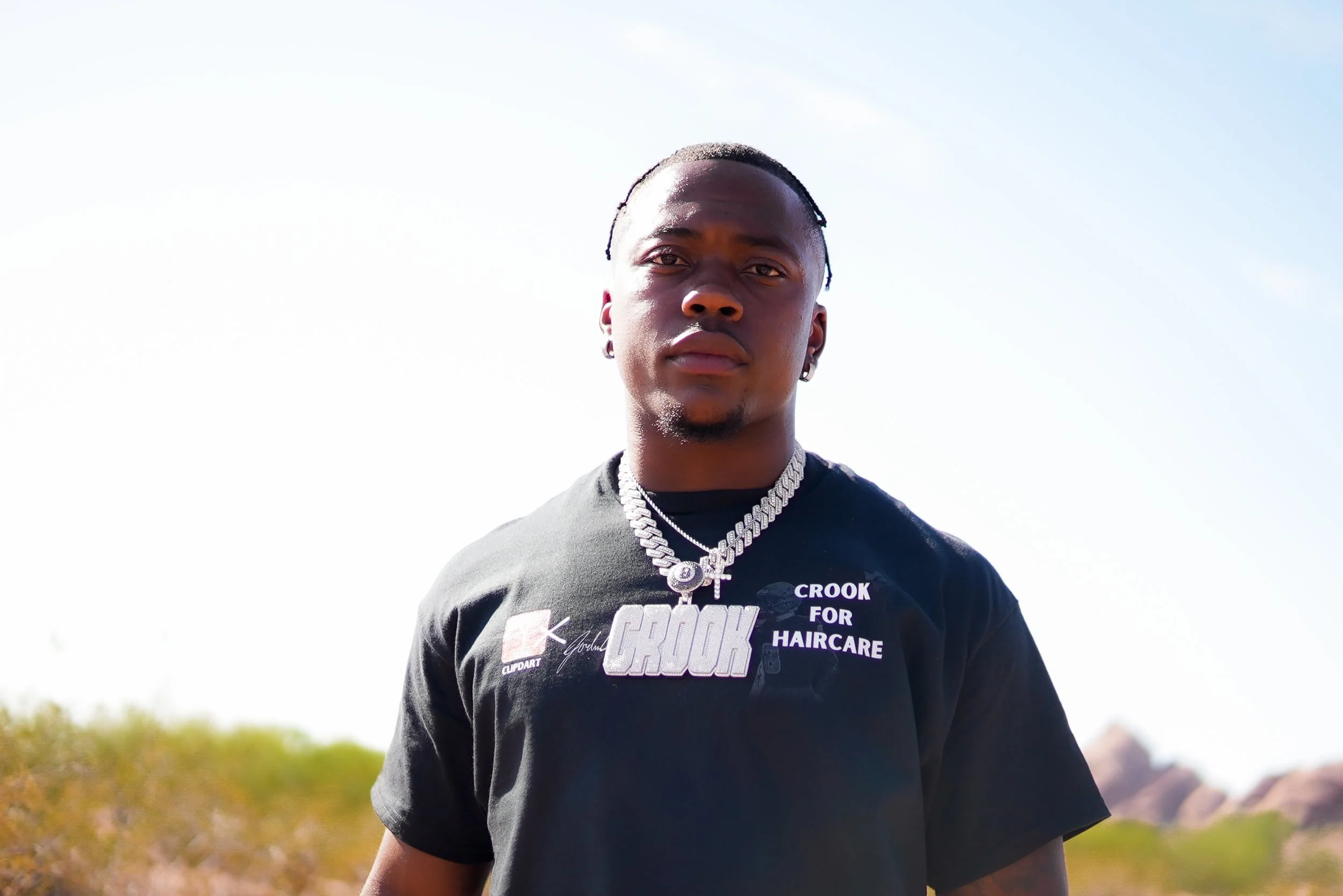 A young man standing outdoors, wearing a black T-shirt with the text 'Crook for Haircare' and a large silver chain with a pendant reading 'Crook.' He has earrings and is looking directly at the camera. The background shows a clear sky and some distan