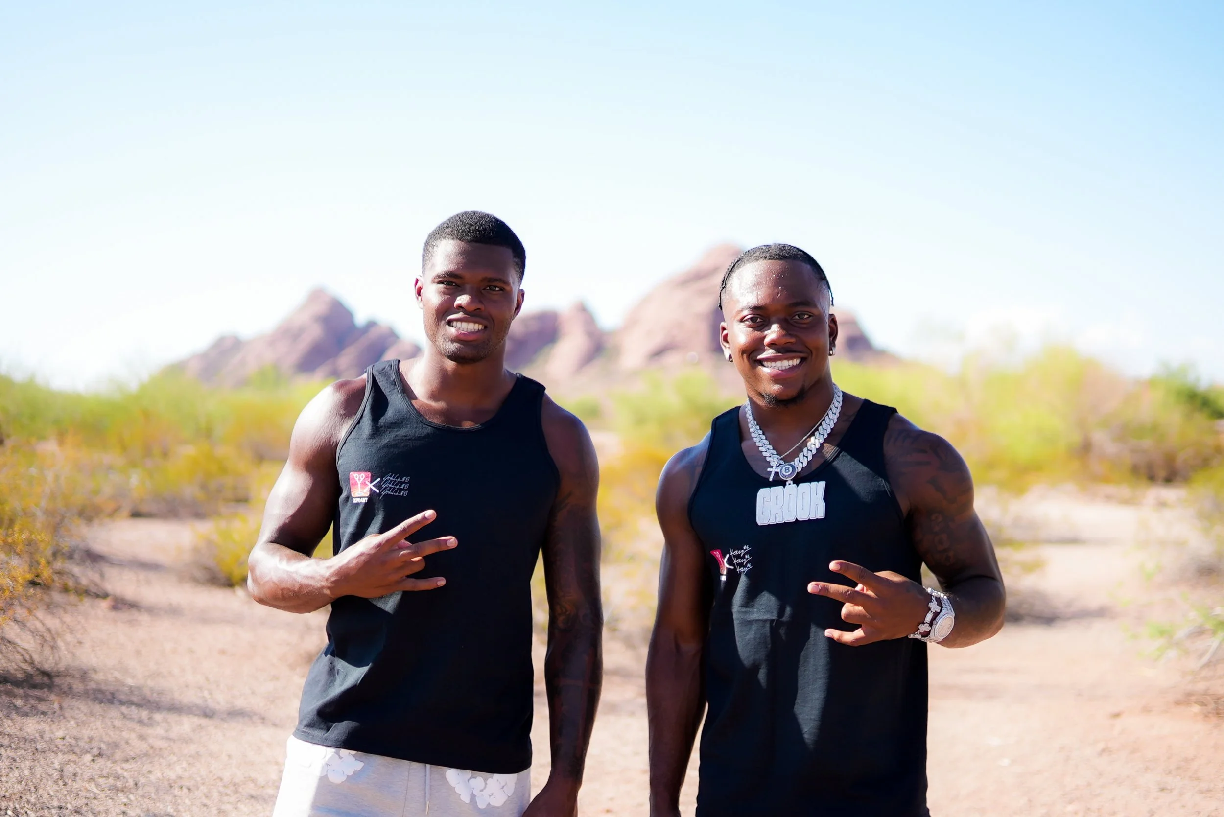 Two young men in sleeveless black shirts posing outdoors on a desert trail with mountains in the background. One is making a peace sign, both smiling.