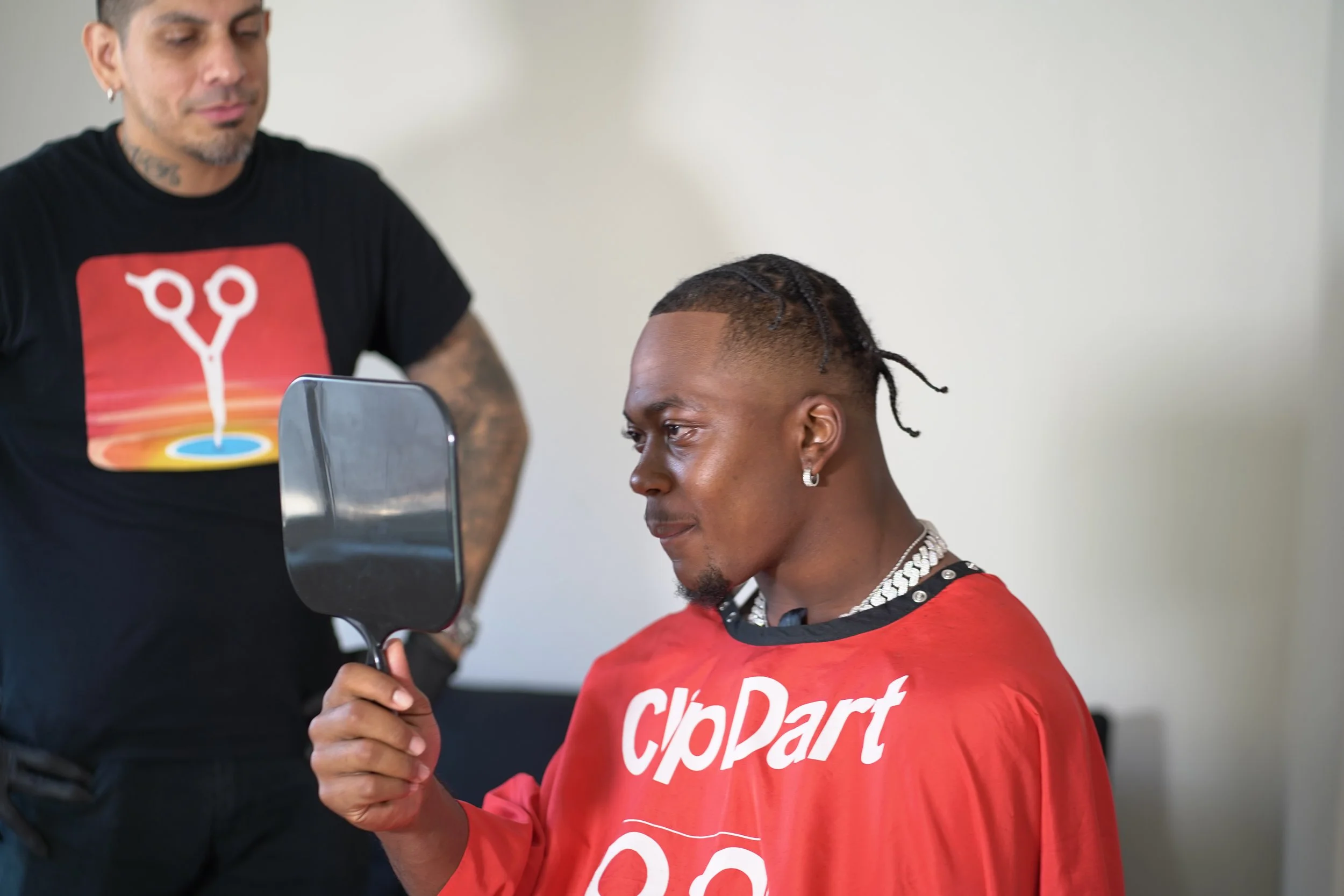 A man wearing a red CliPArt cape looking at his reflection in a mirror held in his hand with another man standing nearby.