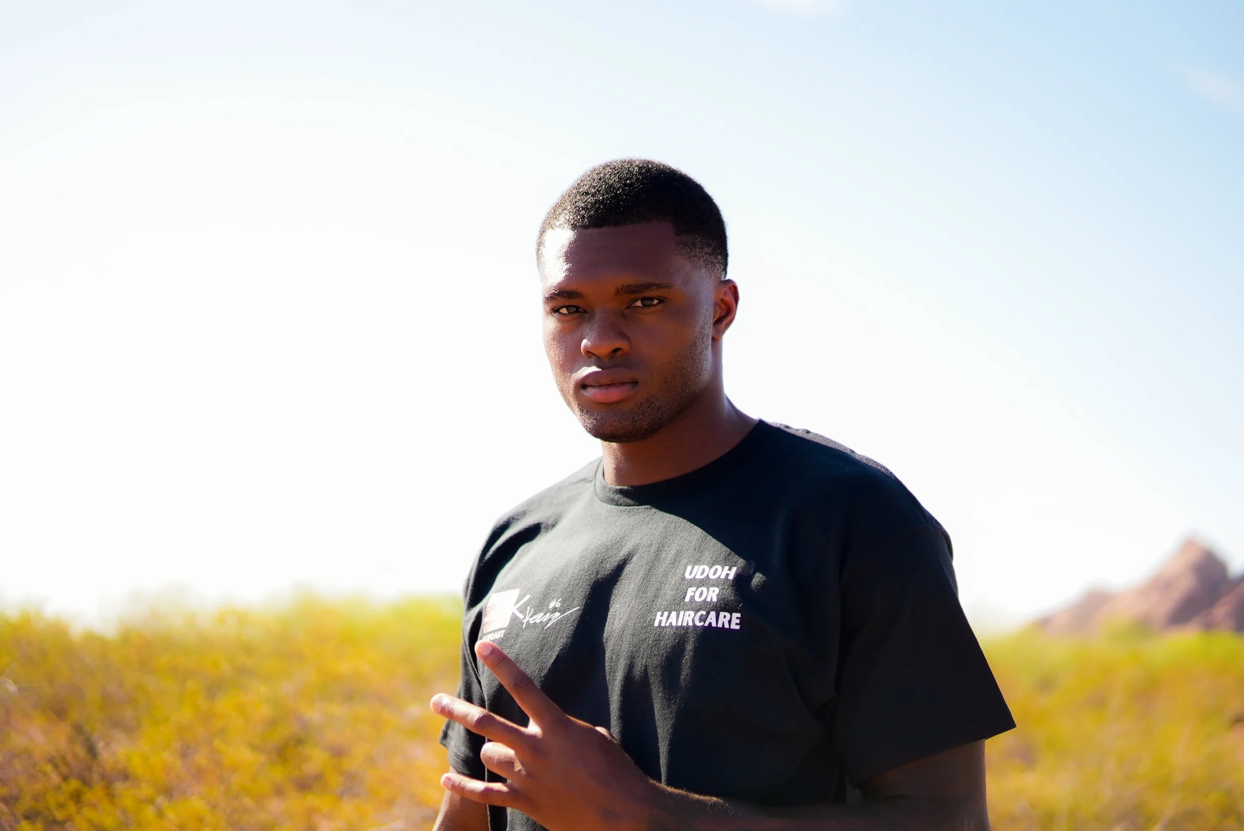 Portrait of a young man with short hair and a serious expression, standing outdoors on a sunny day, in front of a background of bushes and mountains.