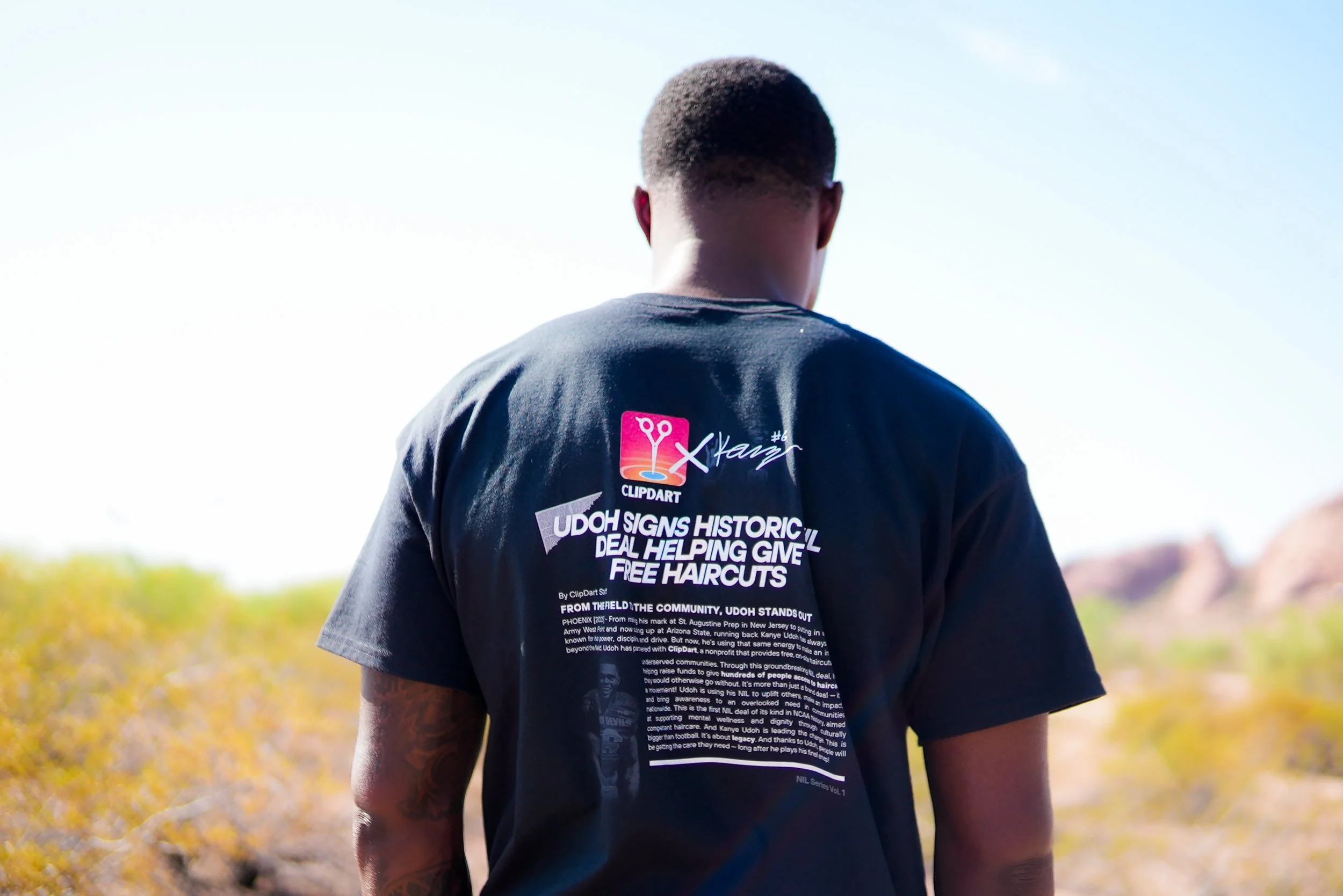Back of a person wearing a black T-shirt with text about a haircuts and community support, standing outdoors with a desert landscape and clear sky in the background.