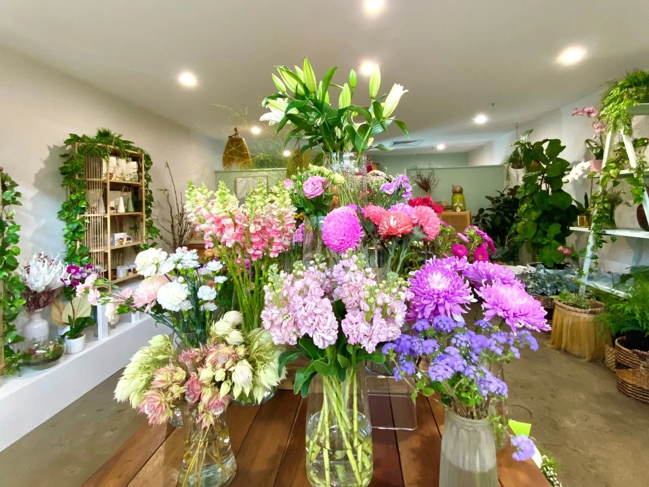 A variety of colorful flowers, including lilies, carnations, and chrysanthemums, arranged in glass vases on a wooden table inside a flower shop.