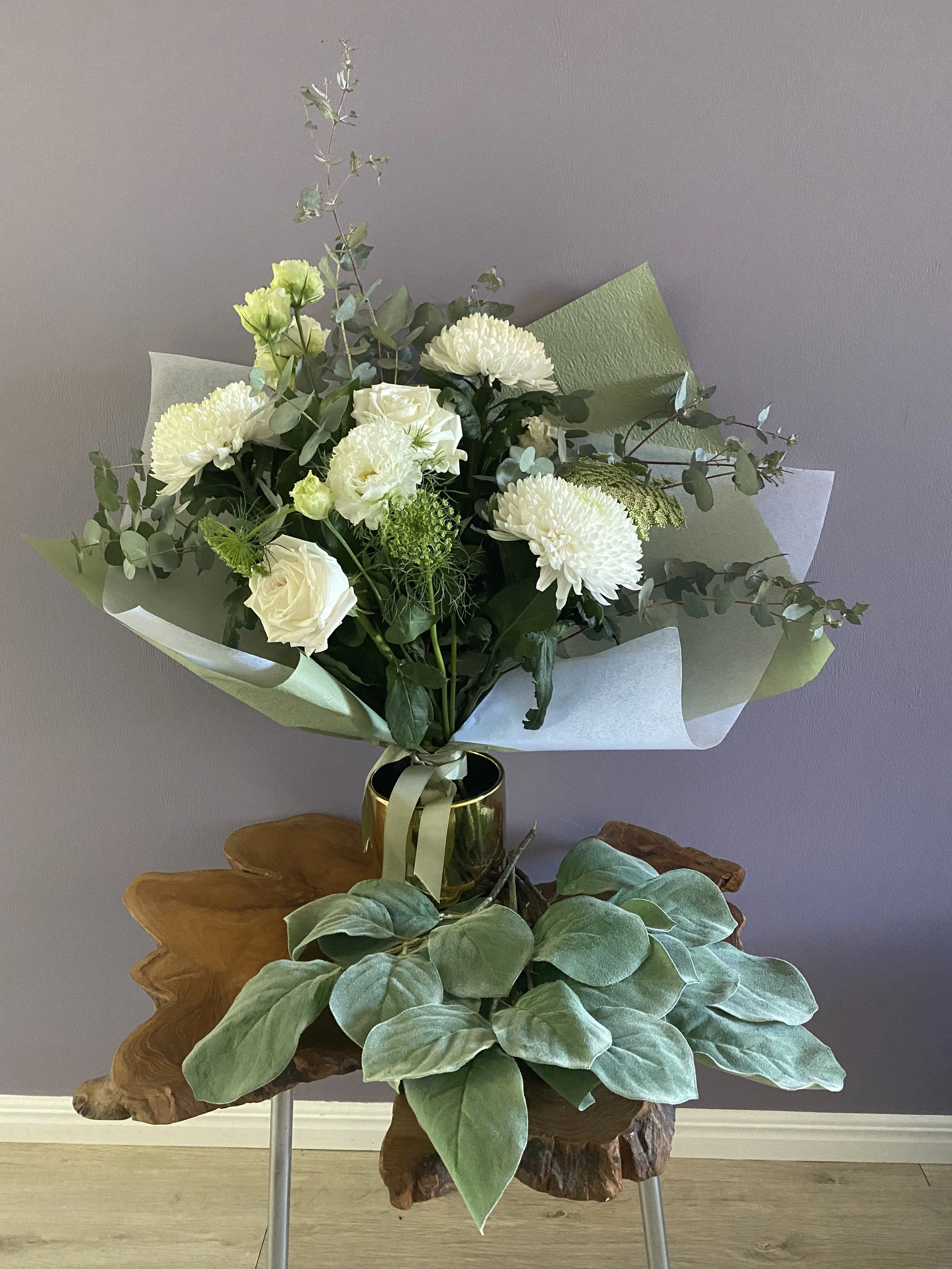 A bouquet of white flowers, including roses, chrysanthemums, and lisianthus, arranged with greenery in a gold vase, placed on a wooden side table with a natural edge, against a gray wall background.